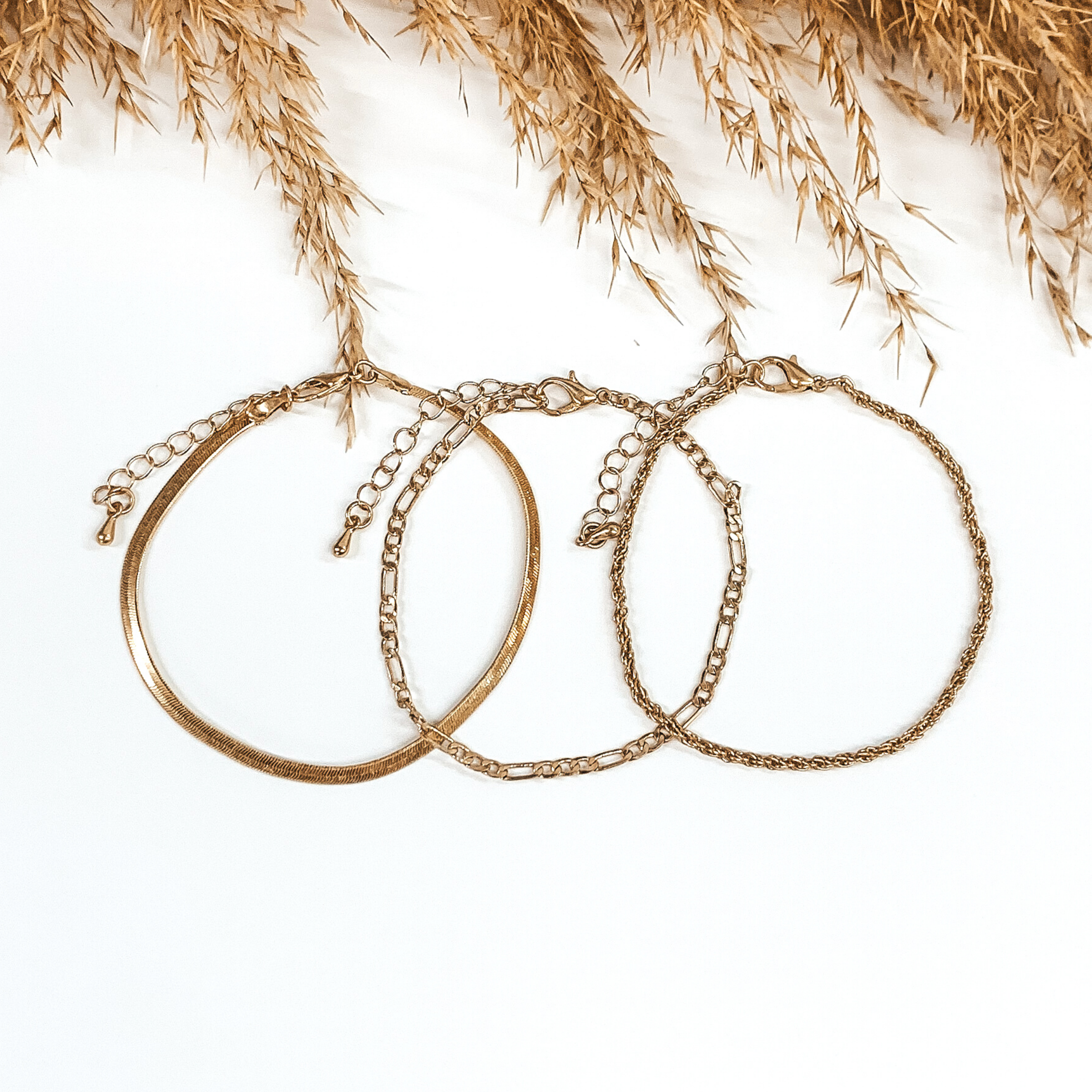 Three gold chained bracelets that are laying on top of each other. There is a snake chain, figaro chain, and a rope chain bracelet. These bracelets are pictured on a white background with tan floral at the top of the picture.