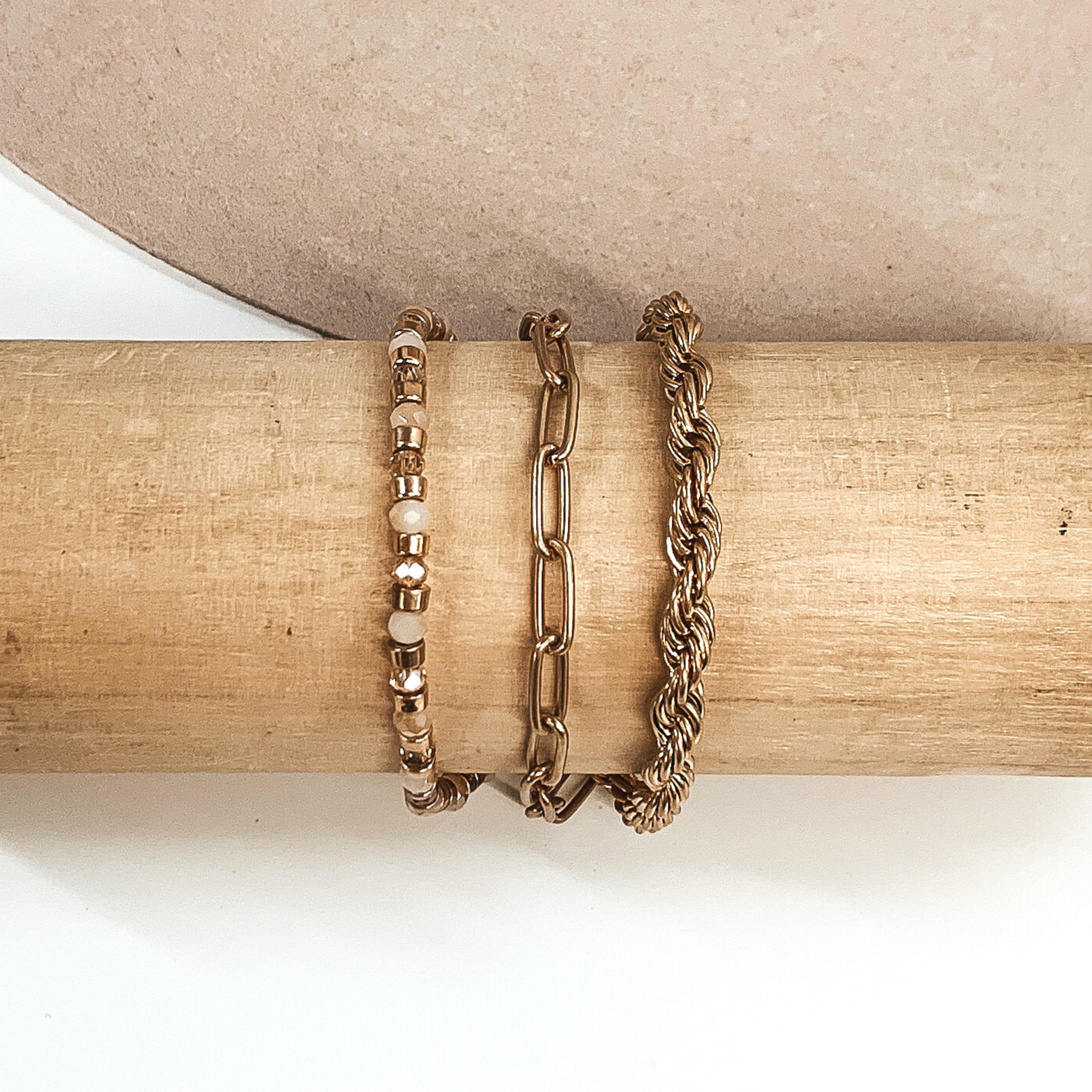This is a gold multi stranded bracelet. This bracelet includes a rope chain, paperclip chain, and a beaded strand. The beaded strand has gold beads, champagne beads, and ivory beads. This bracelet is pictured on a wooden bracelet holder on a white and beige background. 