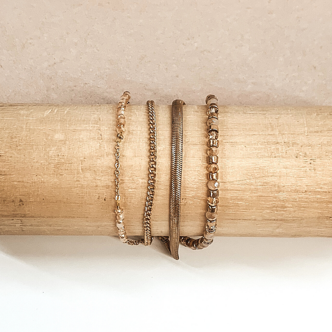 This is a gold multi stranded bracelet. This bracelet includes a snake chain, curb chain, a beaded strand, and a half chain half beaded stranded. The beaded strand has gold beads, champagne beads, and ivory beads. This bracelet is pictured on a wooden bracelet holder on a white and beige background.