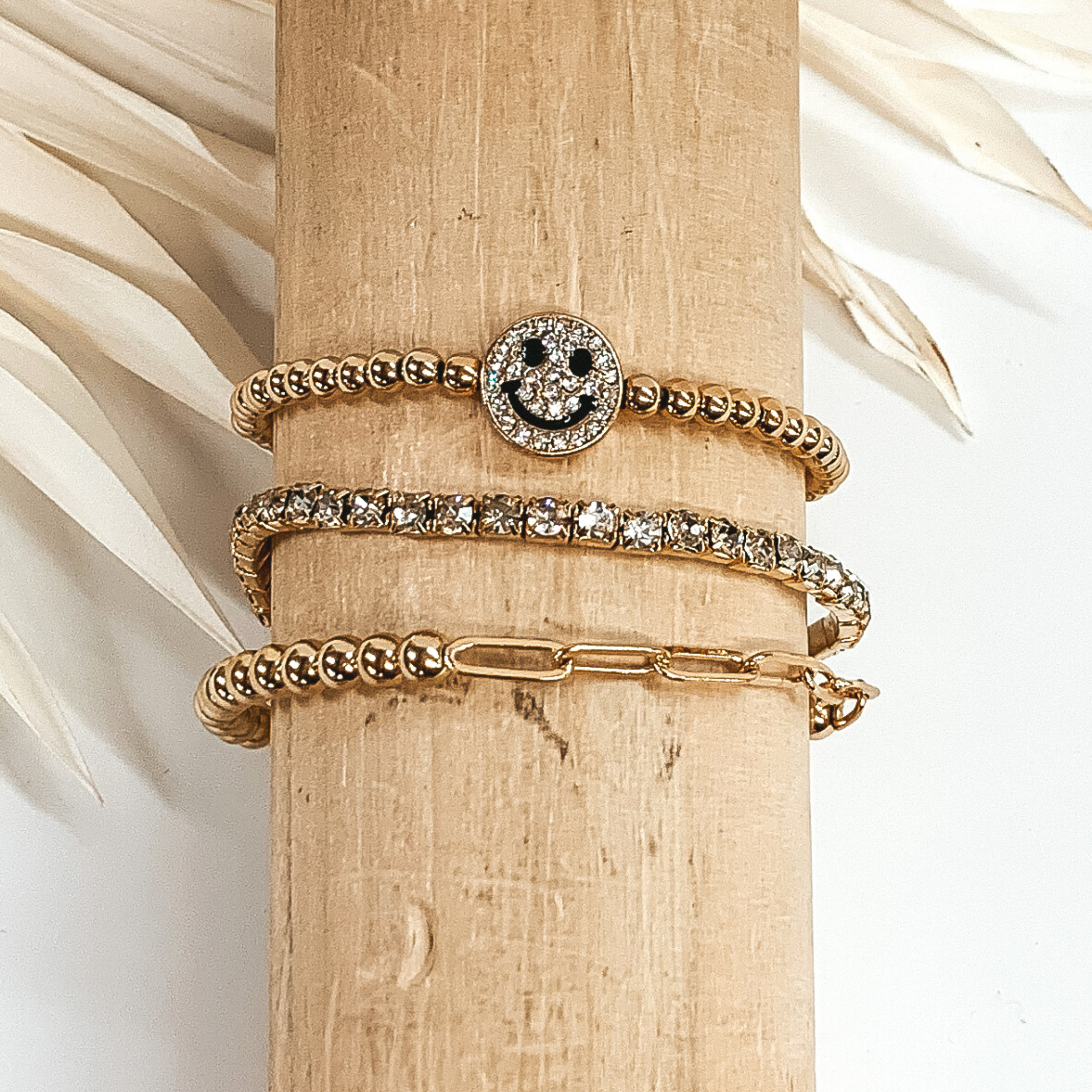 This is a gold bracelet set with a crystal smiley face charm. This set includes a beaded bracelet, a half beaded half chain bracelet, and a clear crystal beaded bracelet. These bracelets are pictured on a wooden bracelet holder on a white background with ivory palm leaves in the background.