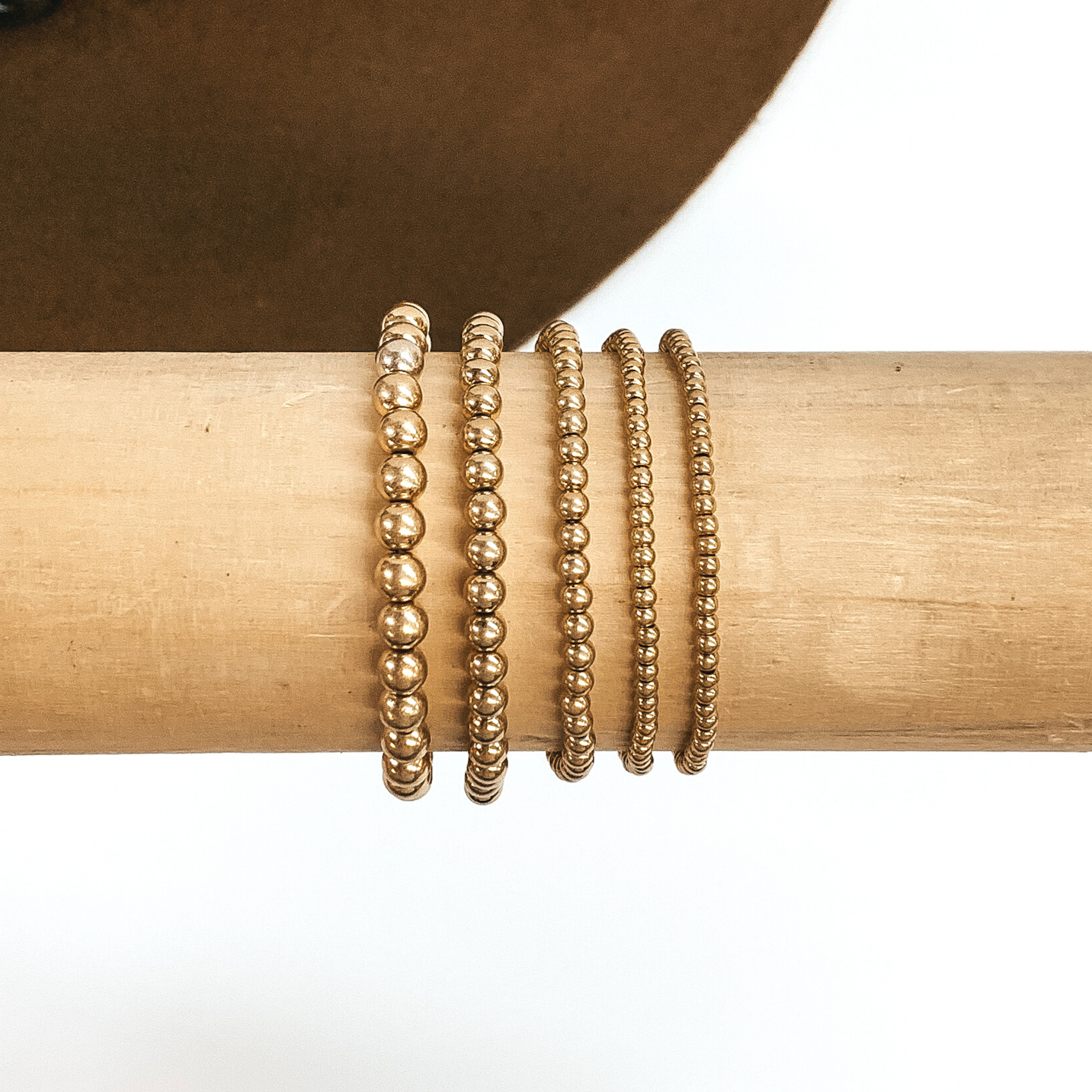 Set of five matte gold beaded bracelets in different sizes. These bracelets are pictured on a wooden bracelet holder on a white and brown background.  