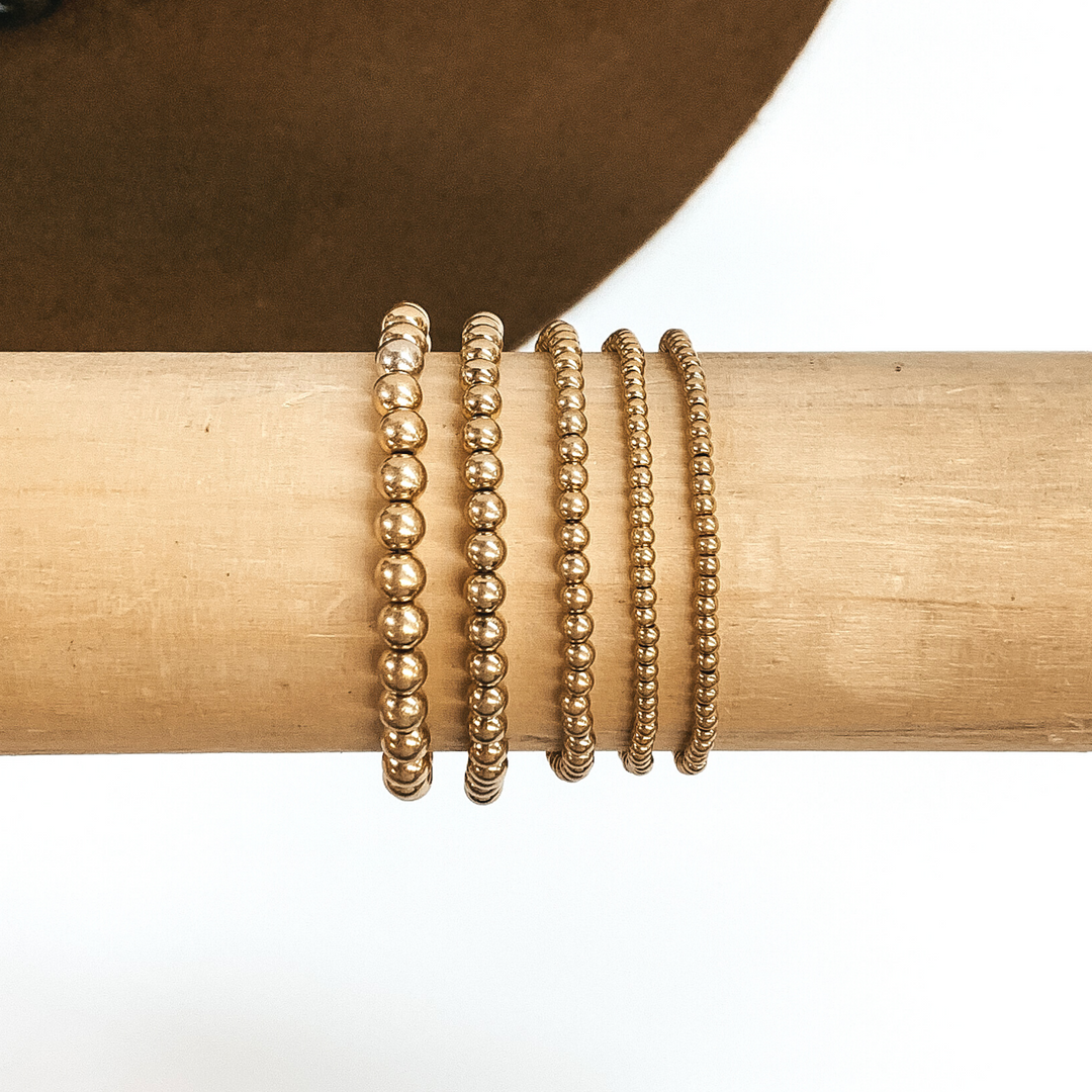 Set of five matte gold beaded bracelets in different sizes. These bracelets are pictured on a wooden bracelet holder on a white and brown background.  