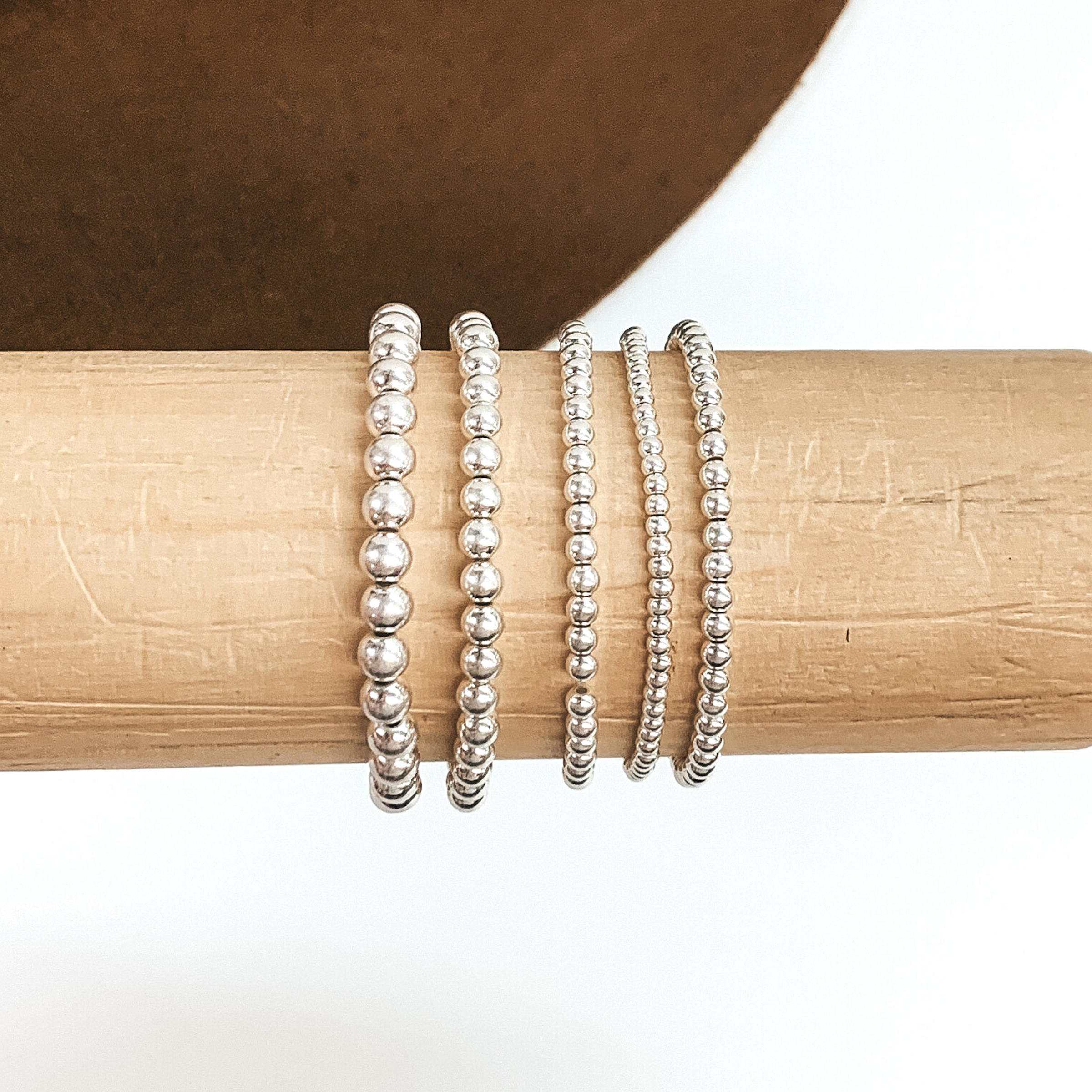 Set of five matte silver beaded bracelets in different sizes. These bracelets are pictured on a wooden bracelet holder on a white and brown background. .