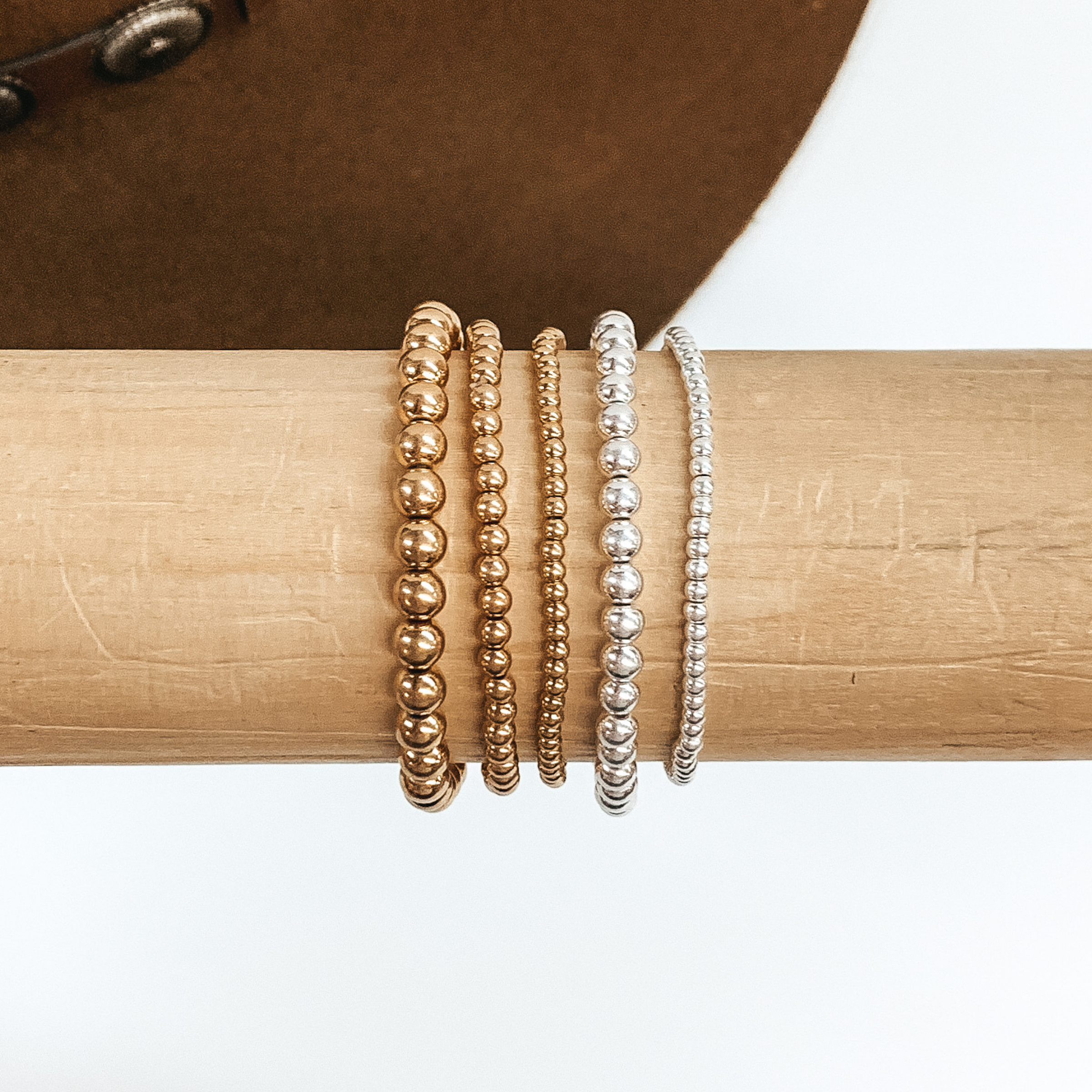 Set of five matte gold and matte silver beaded bracelets in different sizes. These bracelets are pictured on a wooden bracelet holder on a white and brown background.