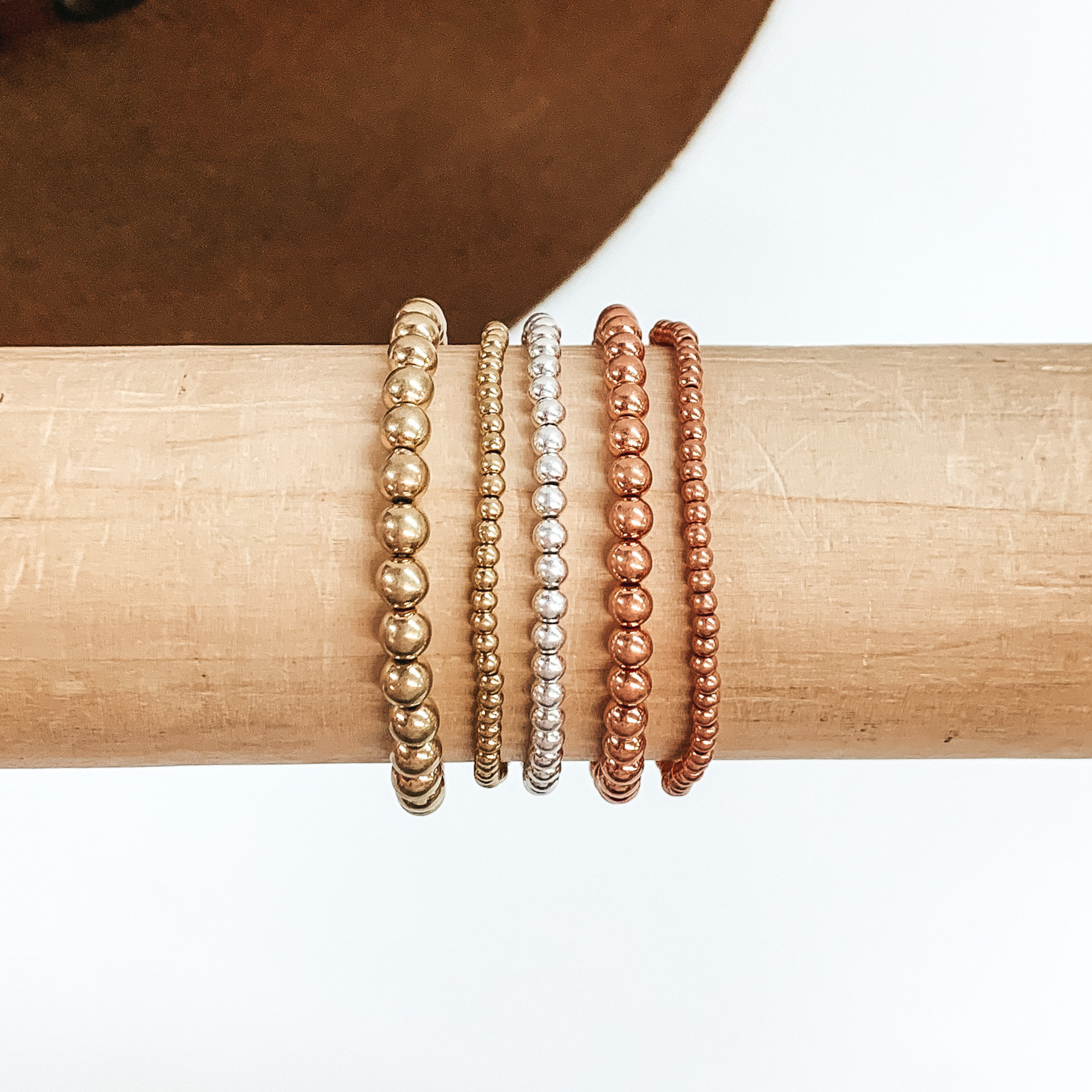 Set of five matte gold, matte silver, and matte copper beaded bracelets in different sizes. These bracelets are pictured on a wooden bracelet holder on a white and brown background. 