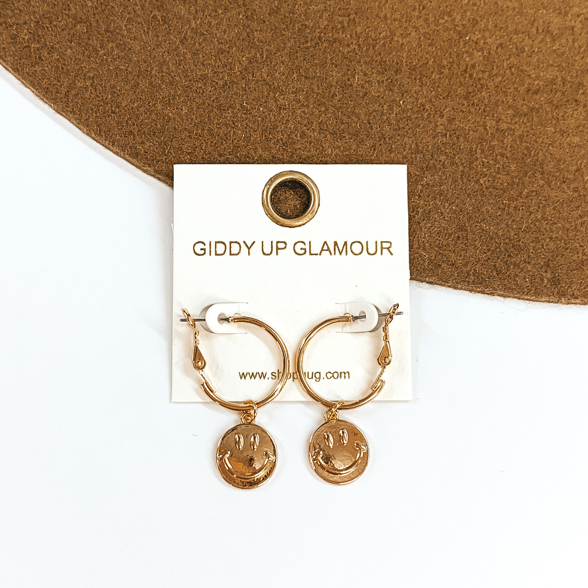 Gold hoop huggies that have hanging gold smiley face charms. These earrings are pictured on a white and brown background. 