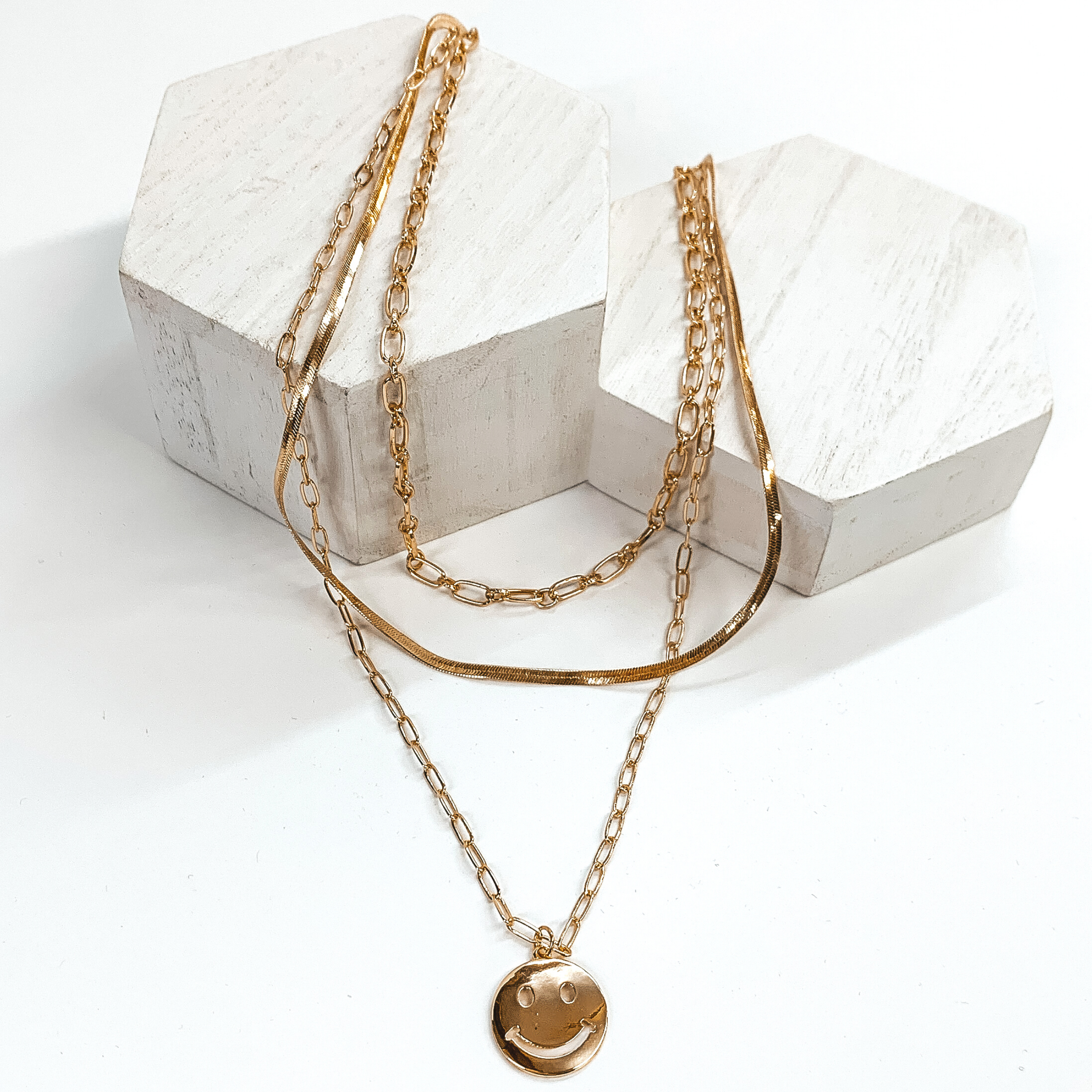 Multi chained gold necklace with the longest strand including a gold smiley face pendant. This necklace is pictured laying on white blocks on a white background. 