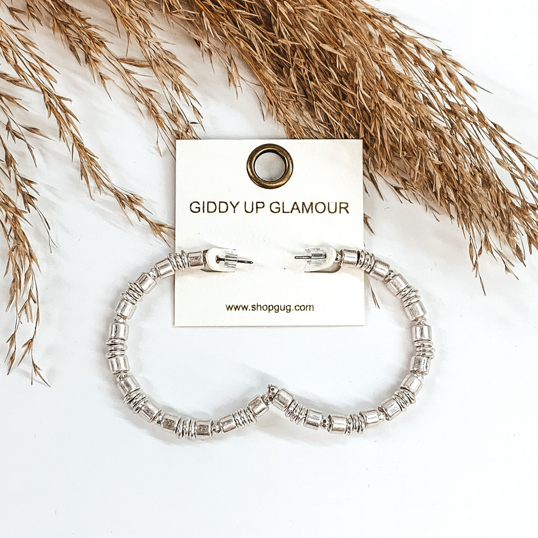 Silver beaded hoop earrings. These earrings include different size beads. These earrings are pictured on a white background with some tan floral at the top of the picture. 