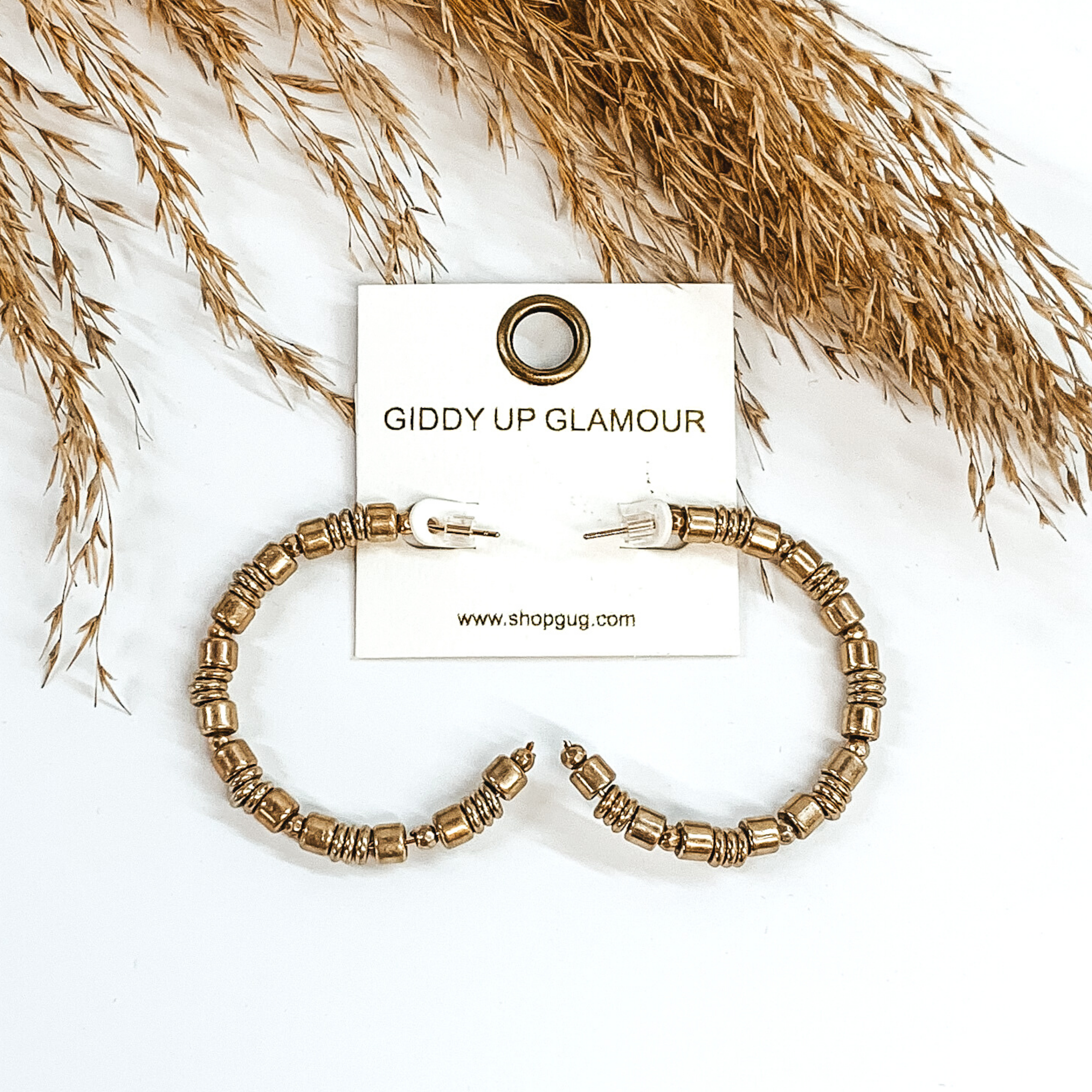 Gold beaded hoop earrings. These earrings include different size beads. These earrings are pictured on a white background with some tan floral at the top of the picture. 