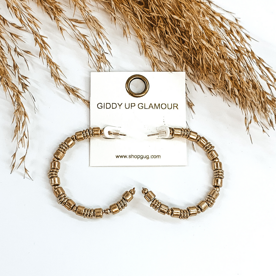 Gold beaded hoop earrings. These earrings include different size beads. These earrings are pictured on a white background with some tan floral at the top of the picture. 