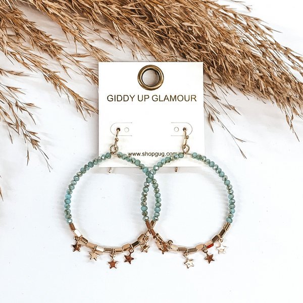 These earrings are a circle drop earrings covered half in turquoise beads and the bottom part has gold beads with tiny gold star charms. These earrings are pictured on a white background with tan floral at the top of the picture. 