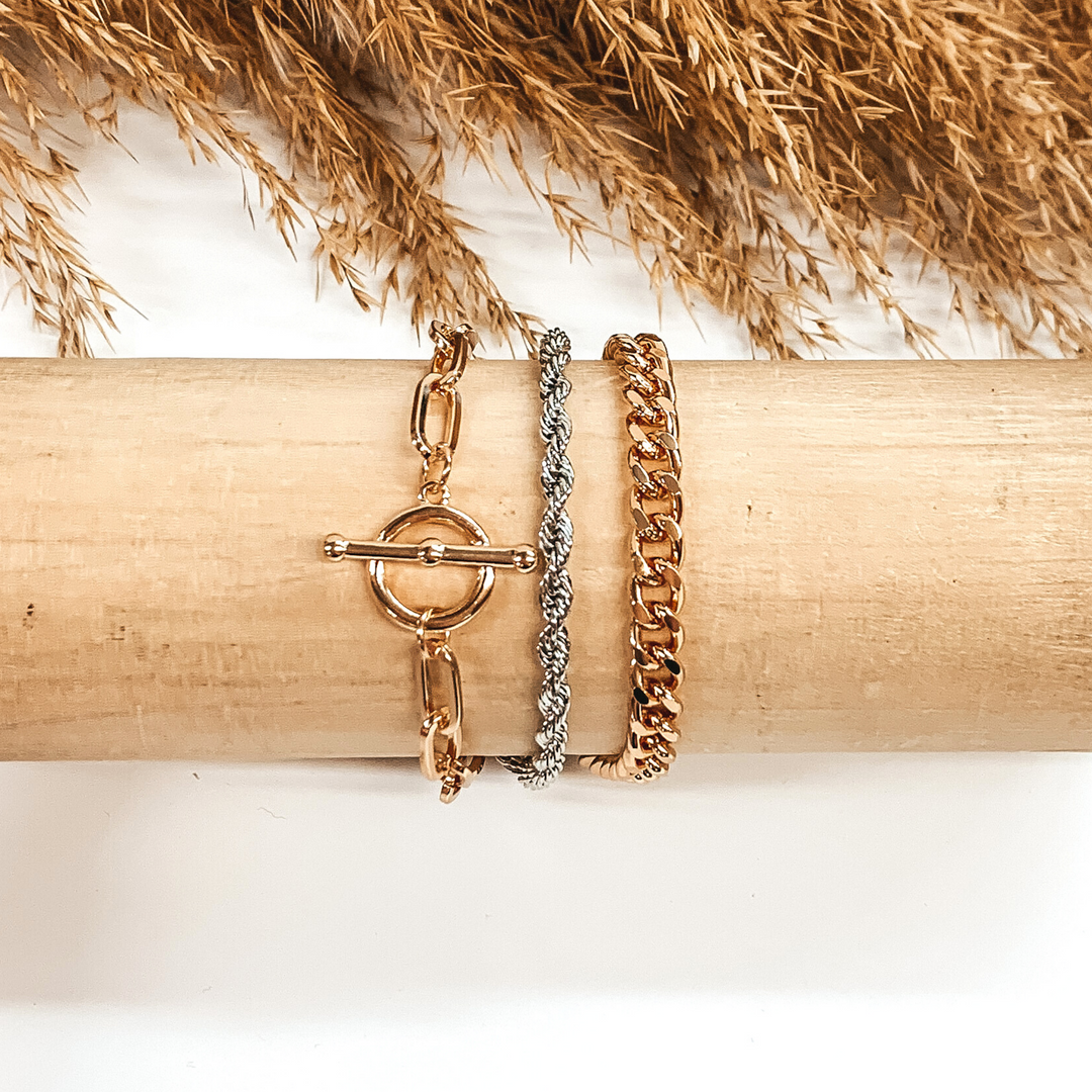 This is magnetic bracelet that includes a gold curb chain, a silver rope chain, and a gold paperclip chain with a toggle clasp pendant. This bracelet is pictured wrapped around a wooden bracelet holder on a white background with tan floral at the top of the picture.