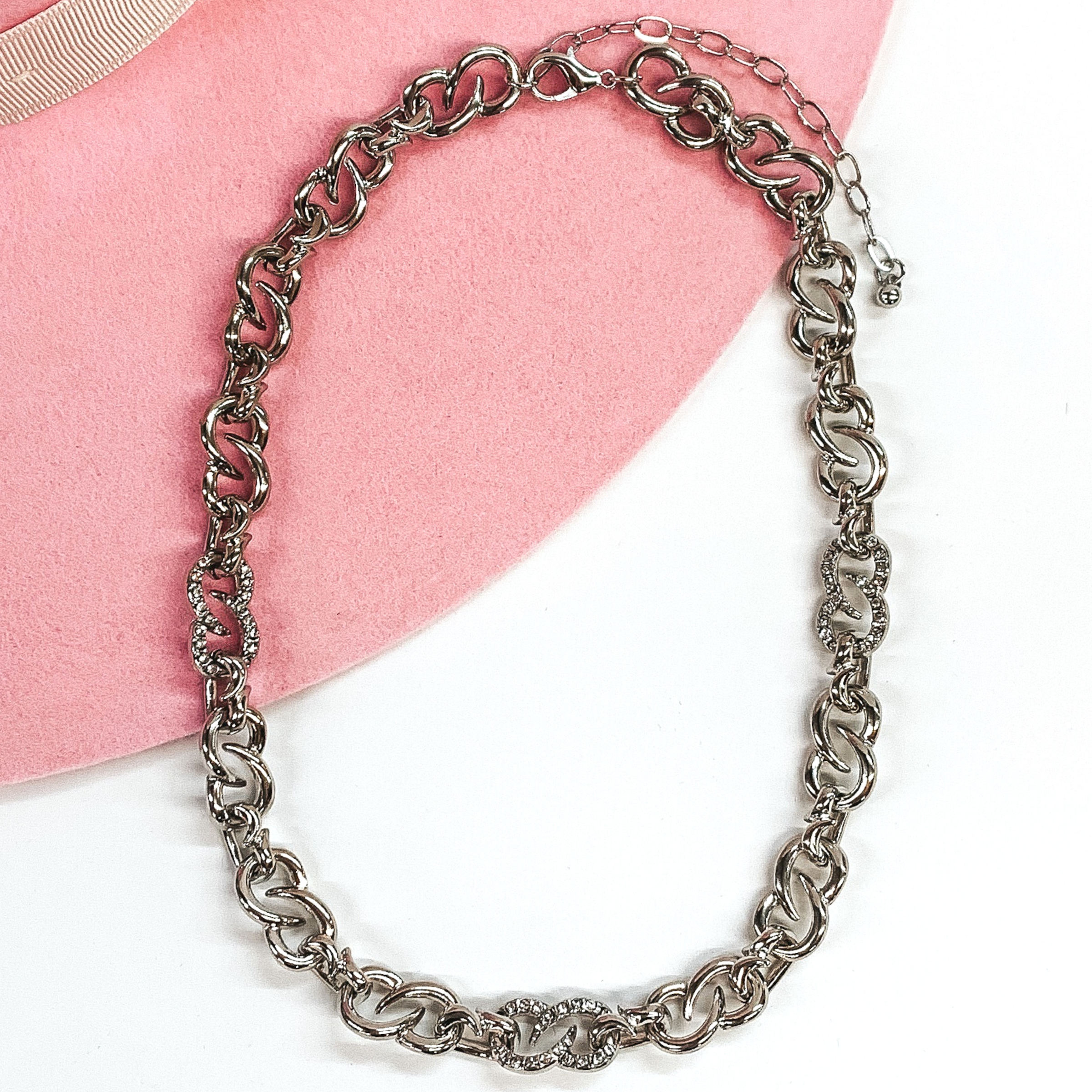 Thick silver infinity chain link necklace with three links that have clear crystals. this necklace is pictured on a white and pink background. 