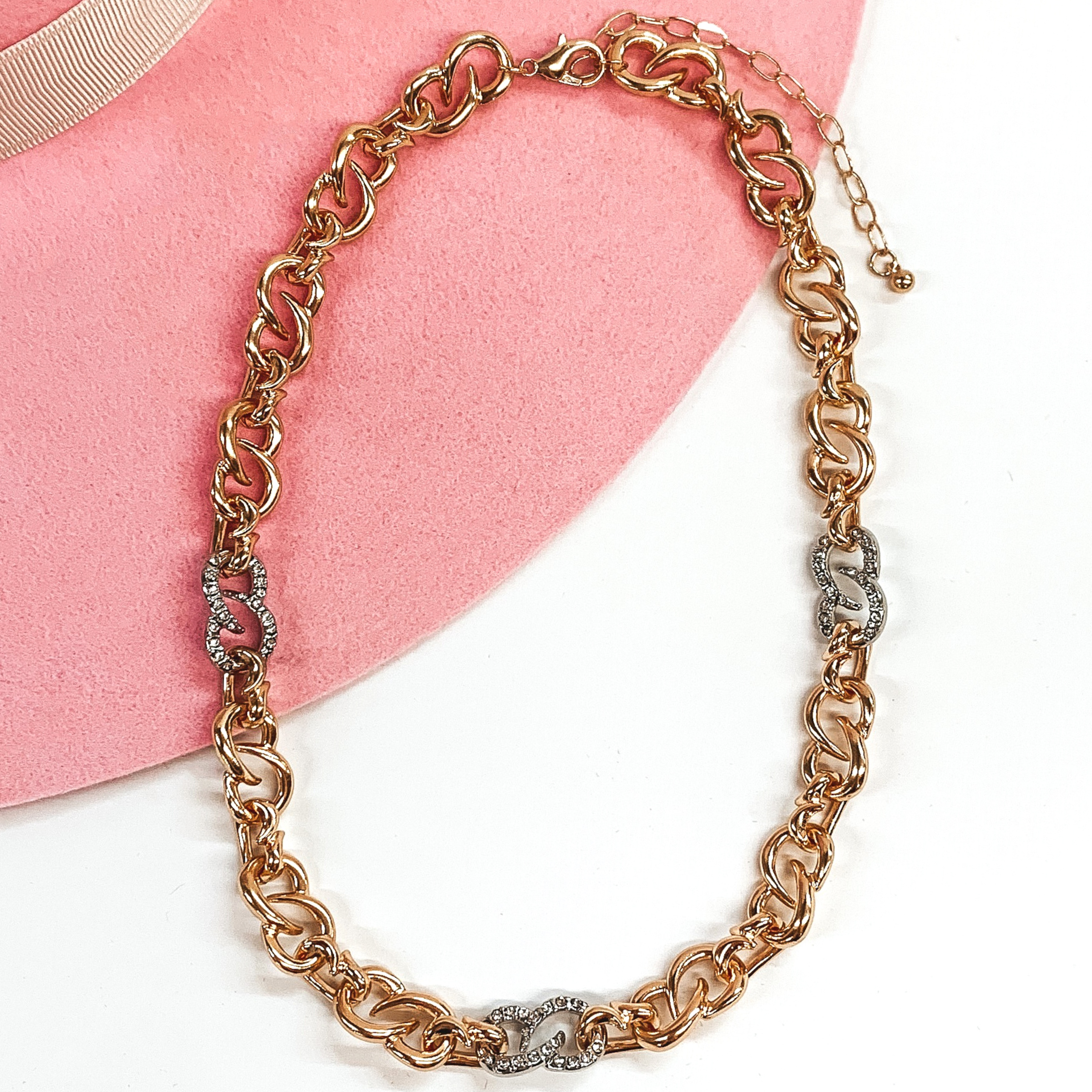 Thick gold infinity chain link necklace with three silver links that have clear crystals. this necklace is pictured on a white and pink background. 