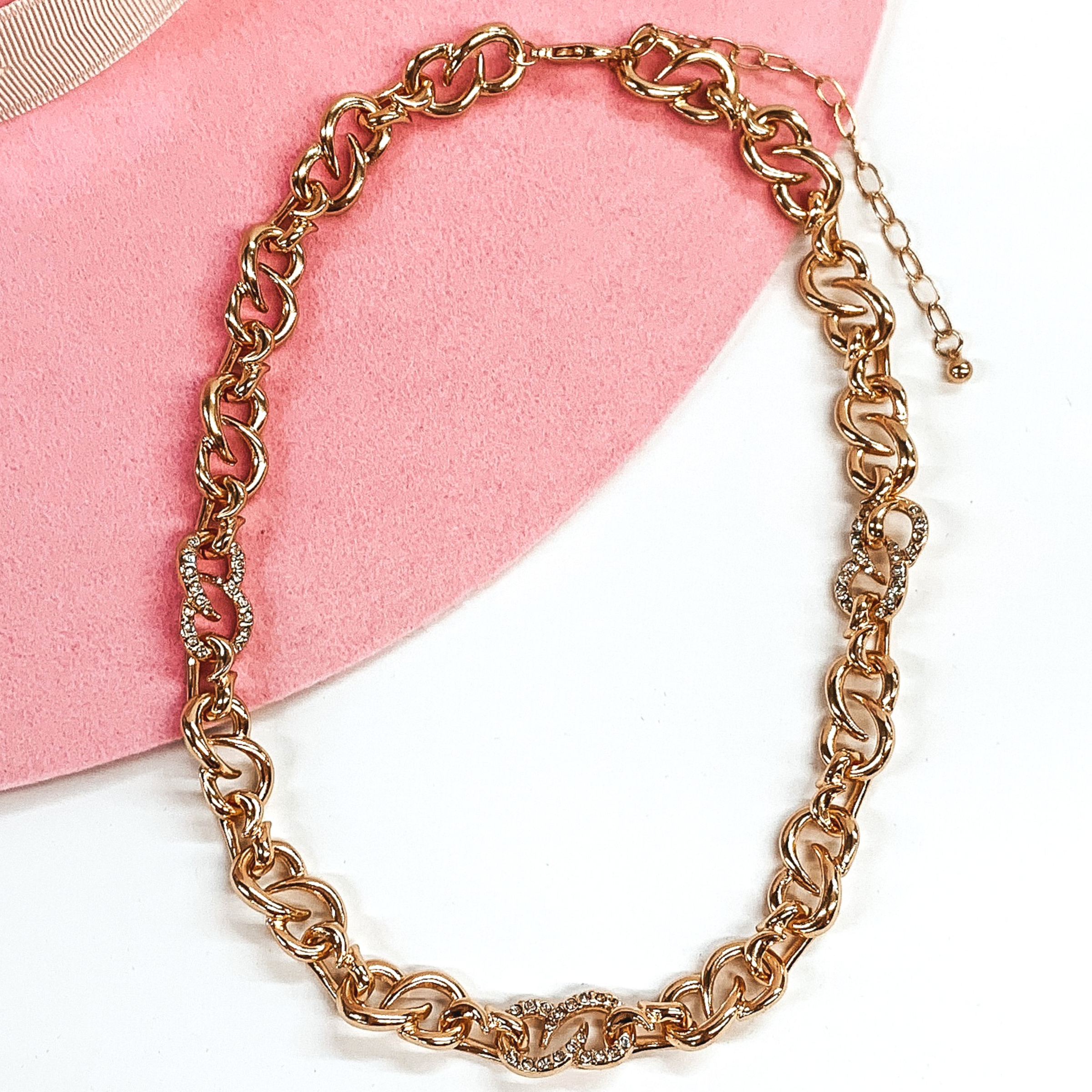 Thick gold infinity chain link necklace with three links that have clear crystals. this necklace is pictured on a white and pink background. 
