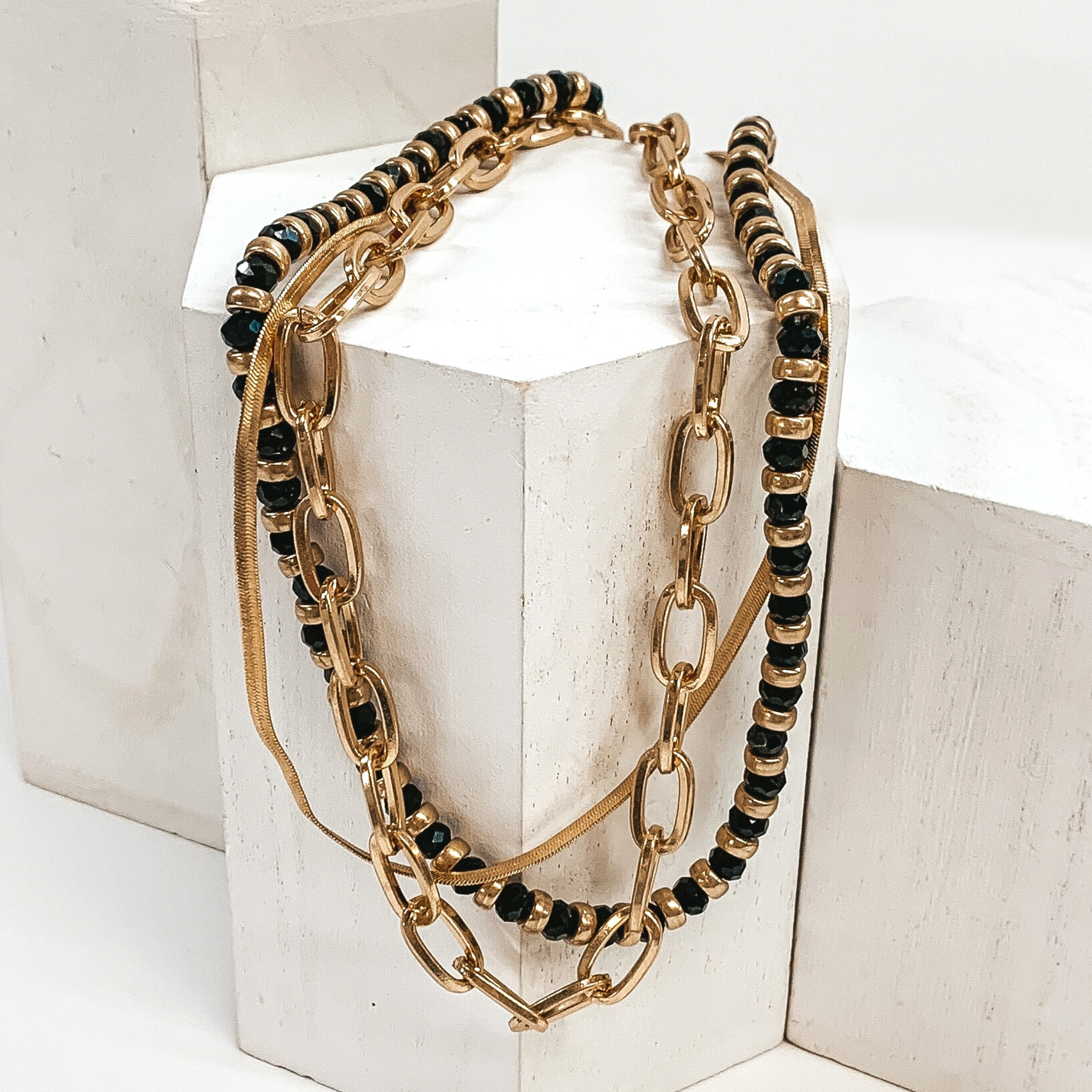 This is a multi layered necklace. One strand is a thick, gold paperclip chain, one is  gold snake chain, and the last strand is a mix of black and gold beads. This necklace is pictured laying on a white block on a white background. 