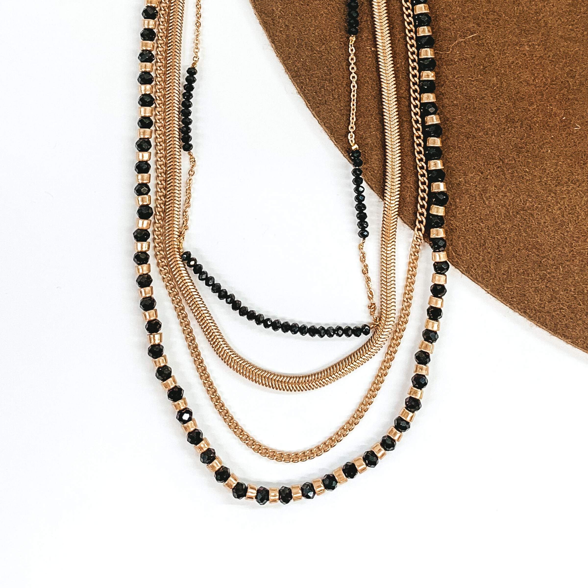 This is a gold multi stranded necklace. This bracelet includes a gold snake chain, a gold curb chain, a black and gold beaded strand, and a half chain half black beaded stranded. This necklace is pictured on a white and brown background.