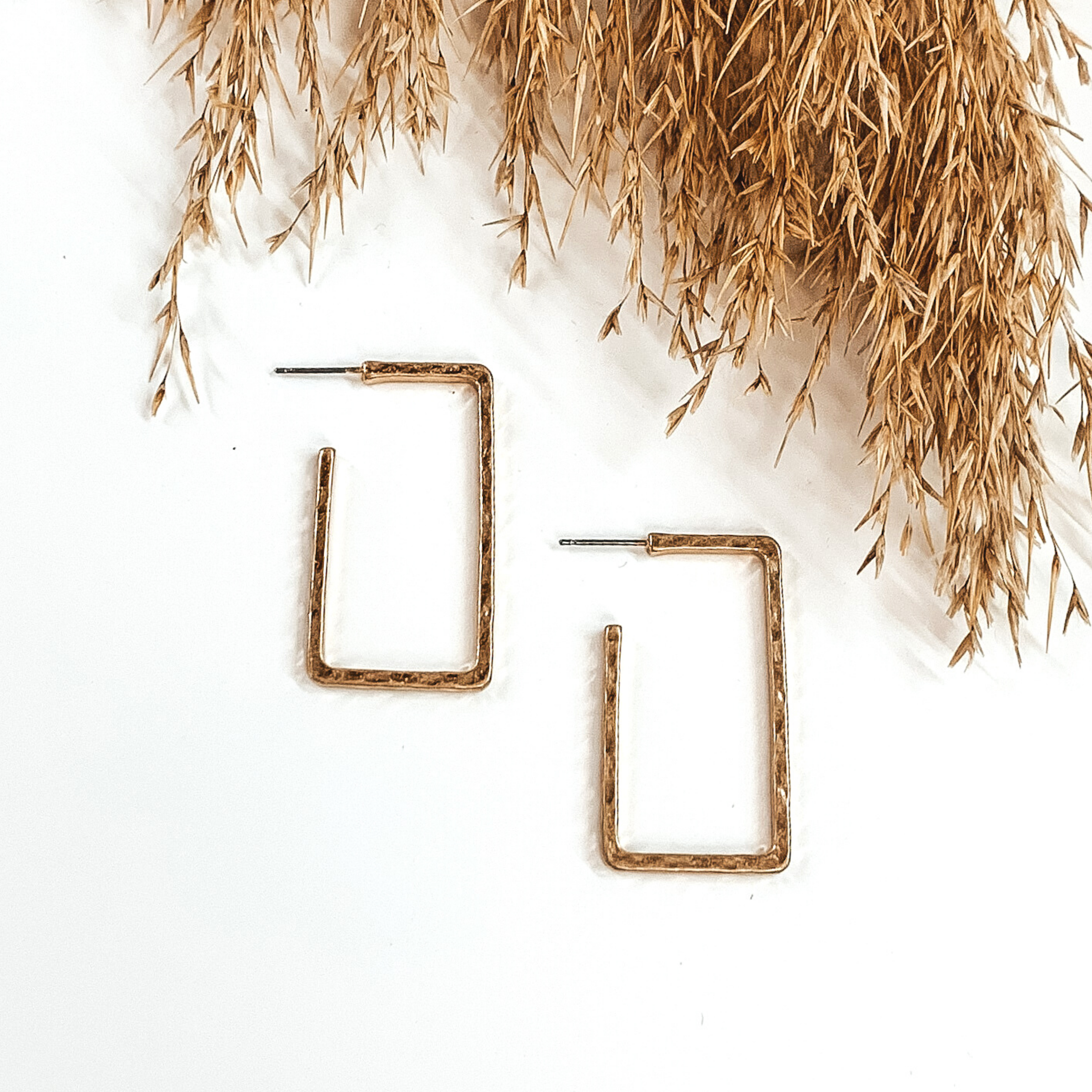 Gold rectangle hoops with a hammered look. These hoops are pictured on a white background with tan floral at the top of the picture. 
