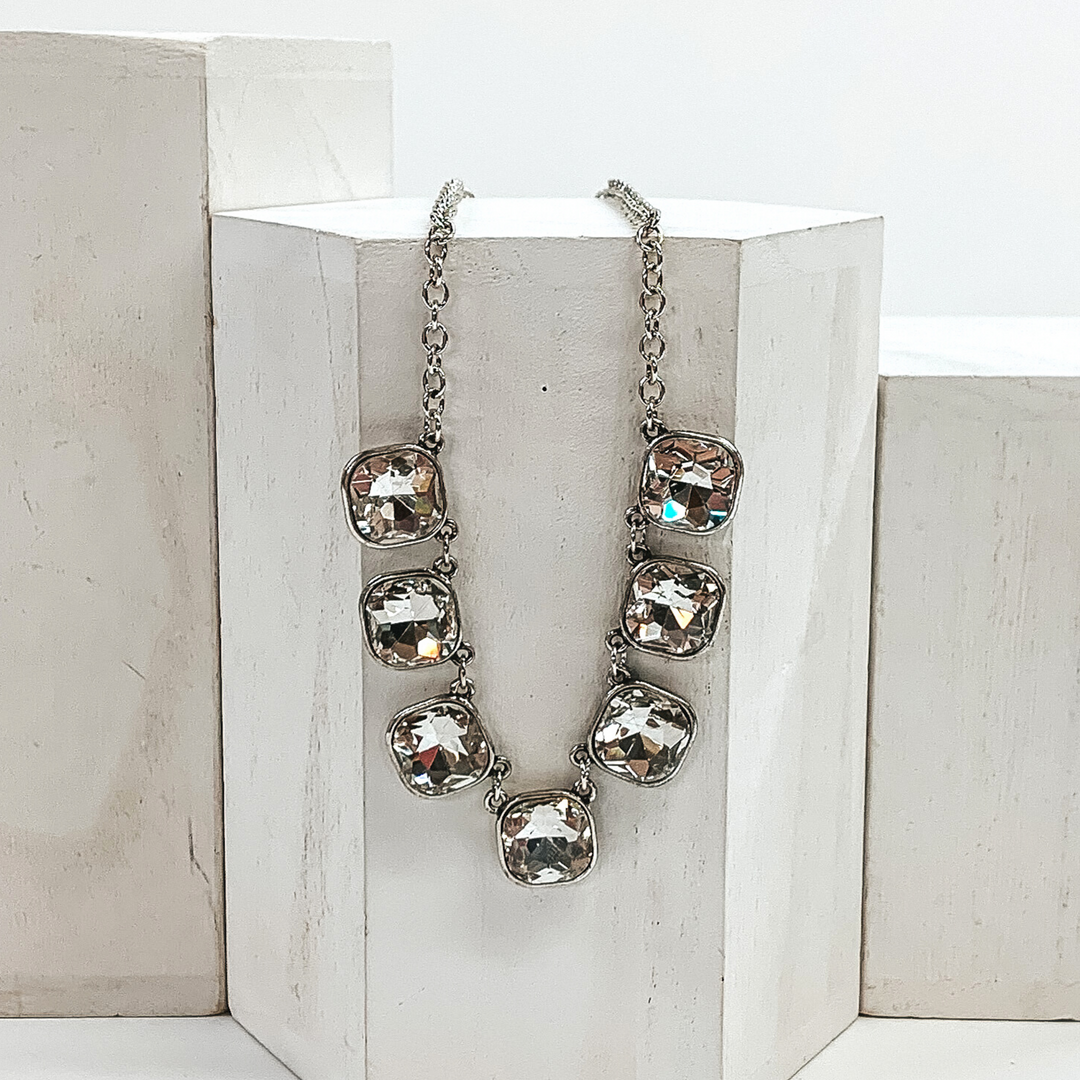 This is a silver chained necklace with seven clear cushion cut crystals. This necklace is pictured laying on a white block on a white background