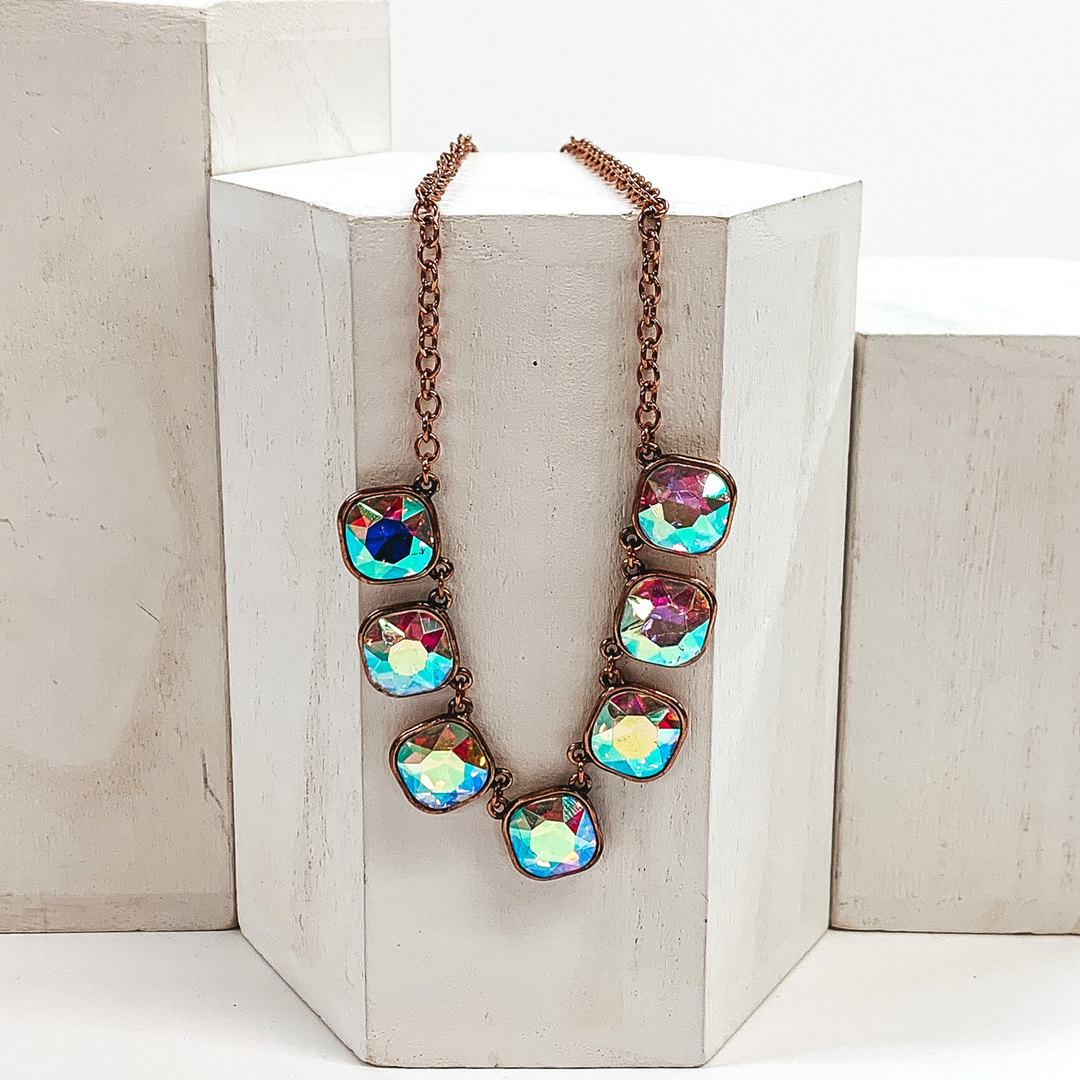 This is a copper chained necklace with seven AB cushion cut crystals. This necklace is pictured laying on a white block on a white background.