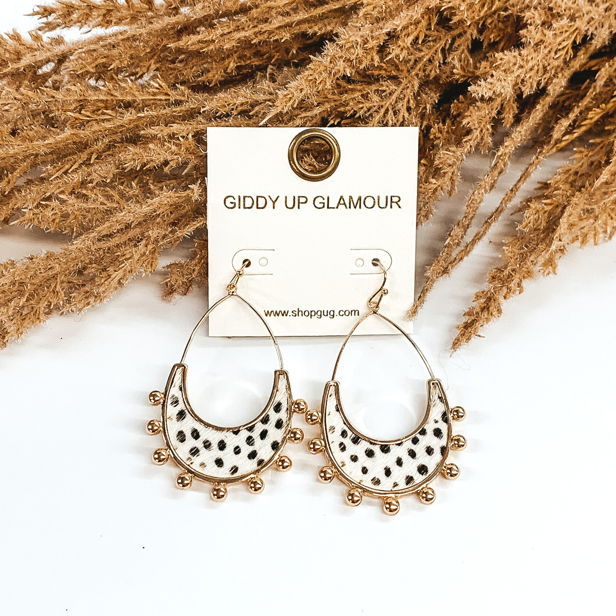 Gold open teardrop earrings with gold beads outlining the bottom of the earrings. There is a white genuine cow hide inlay with black dotted print. These earrings are pictured on a white background and laying against some tan floral. 