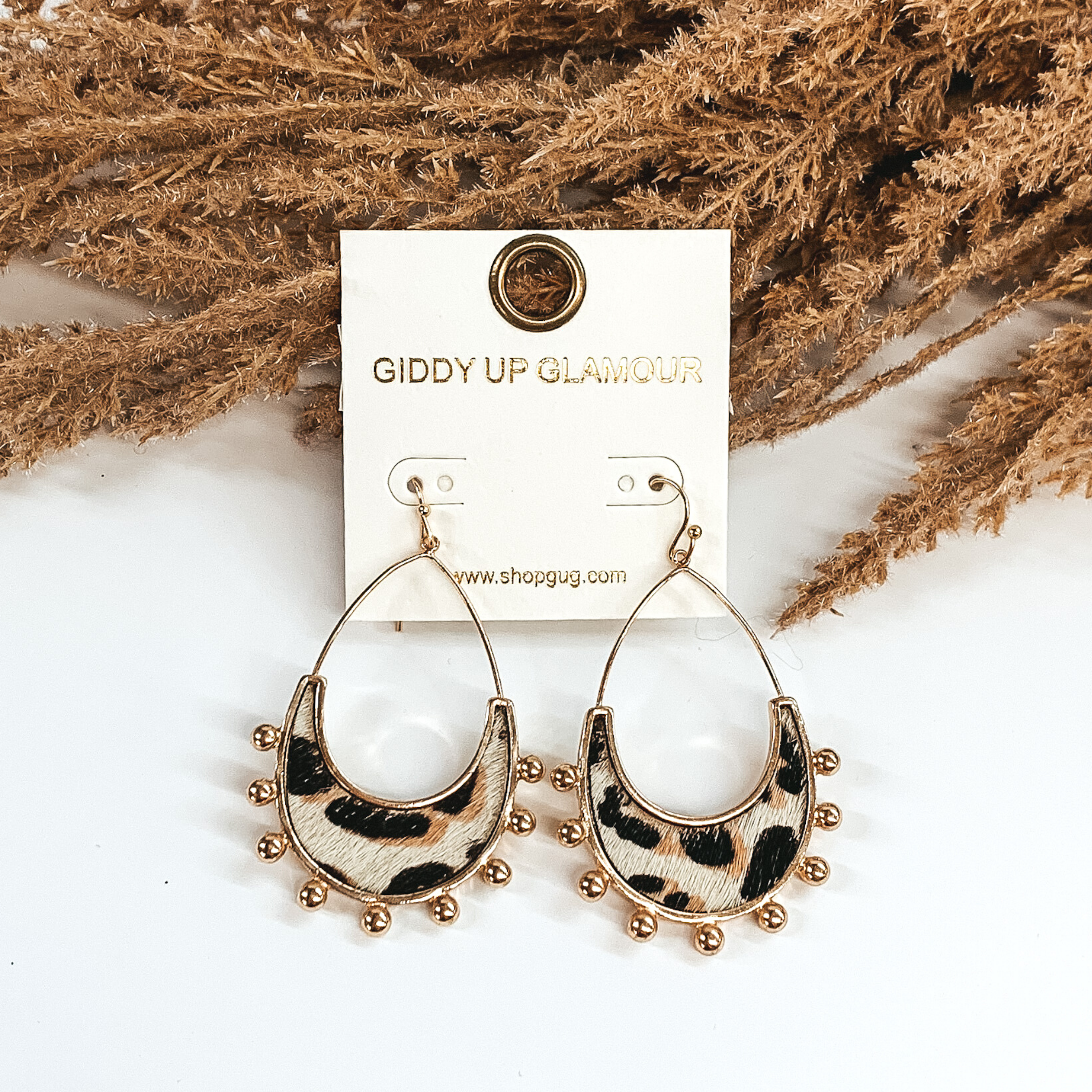 Gold open teardrop earrings with gold beads outlining the bottom of the earrings. There is a white genuine cow hide inlay with a black and brown animal print. These earrings are pictured on a white background and laying against some tan floral. 