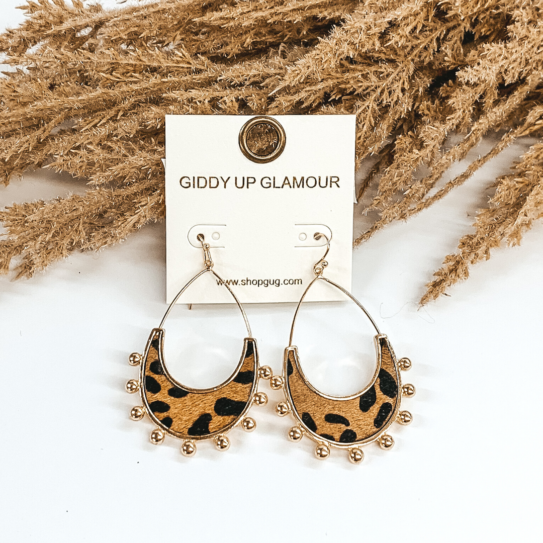 Gold open teardrop earrings with gold beads outlining the bottom of the earrings. There is a brown genuine cow hide inlay with a black animal print. These earrings are pictured on a white background and laying against some tan floral. 