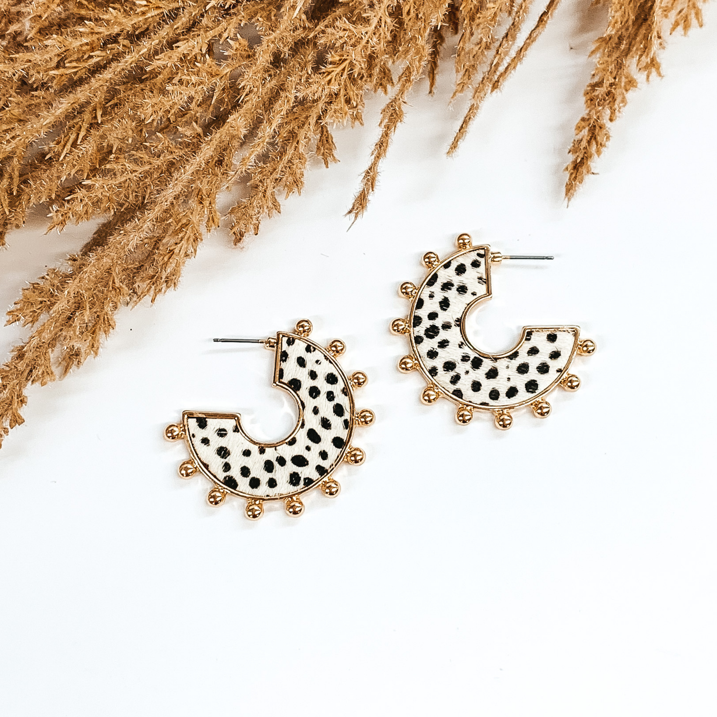 Flat hoops with white animal hide inlay with black dotted pattern. These hoops have gold beads on the edge of the earrings. These earrings are pictured o  white background with tan floral at the top of the page. 