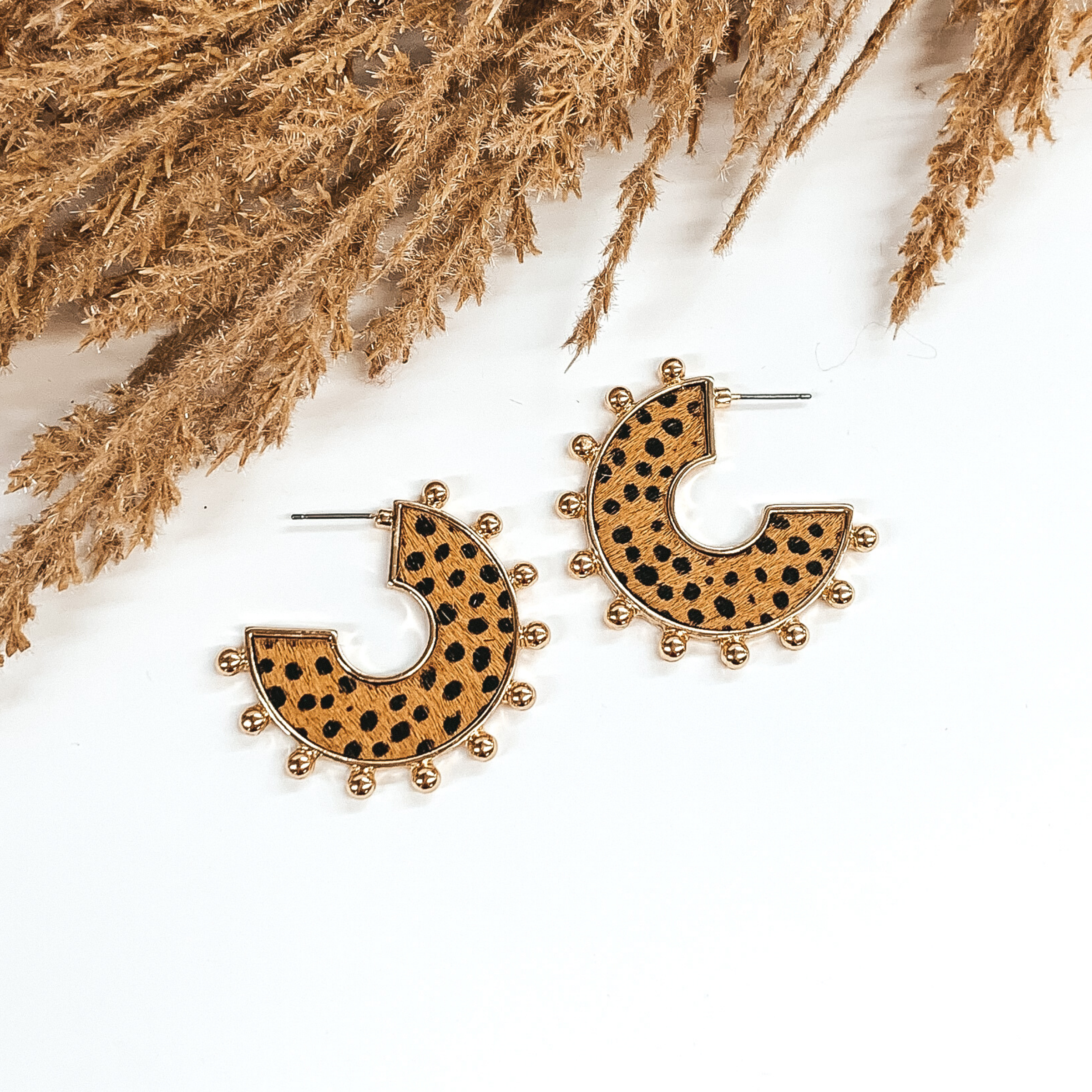 Flat hoops with brown animal hide inlay with black dotted pattern. These hoops have gold beads on the edge of the earrings. These earrings are pictured o  white background with tan floral at the top of the page. 