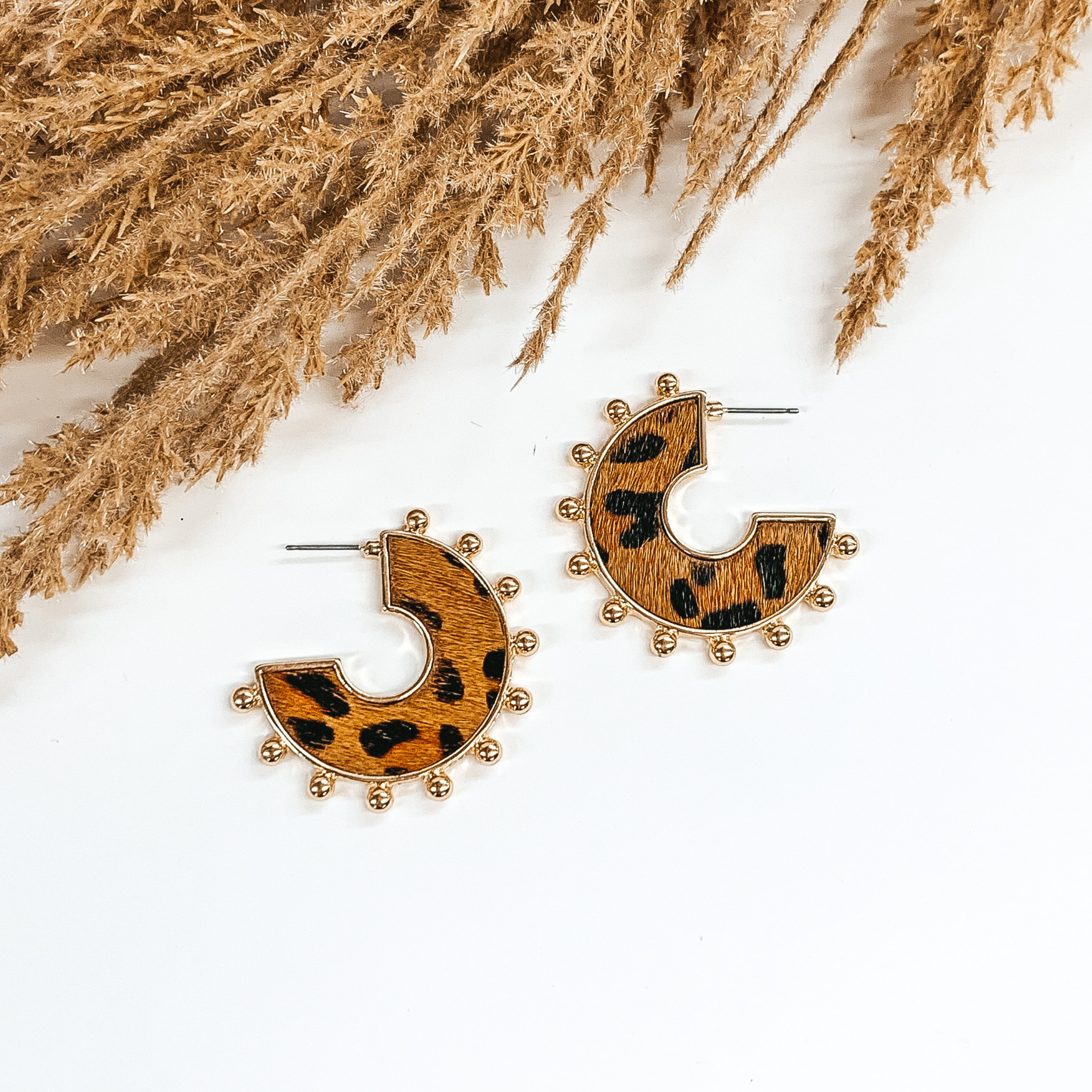 Flat hoops with brown animal hide inlay with black animal print pattern. These hoops have gold beads on the edge of the earrings. These earrings are pictured o  white background with tan floral at the top of the page. 