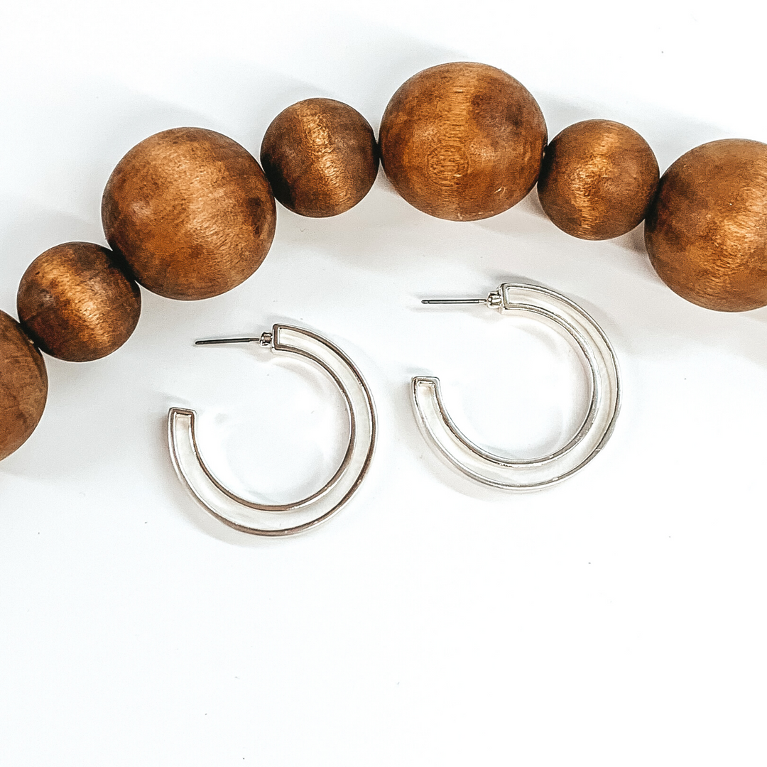 Laying on a white background are two silver hoop earrings. They are a circle double layered hoop earrings. There are brown decor beads above the earrings. 