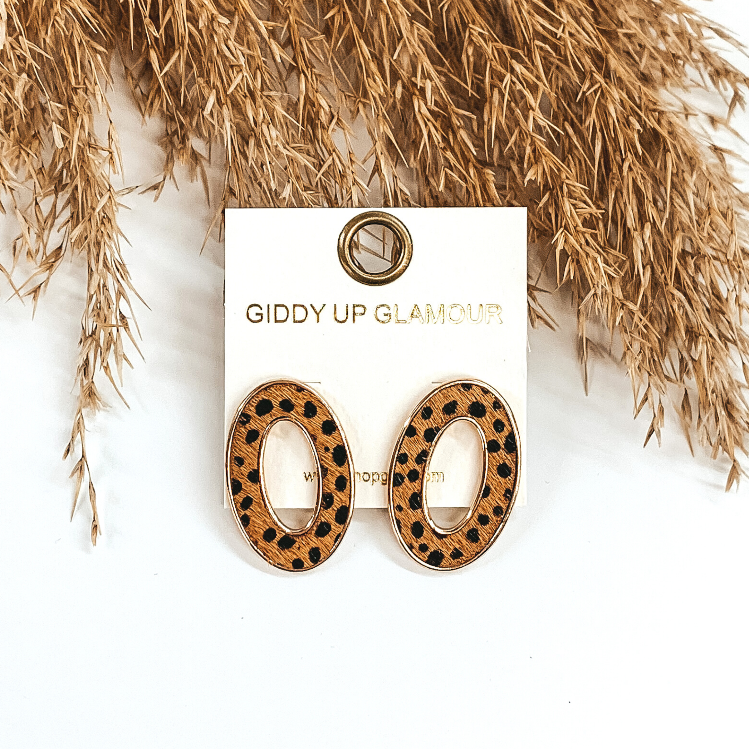 Gold, open, oval stud earrings with a brown hide inlay that has a black dotted print. These earrings are pictured laying on top of tan floral on a white background. 