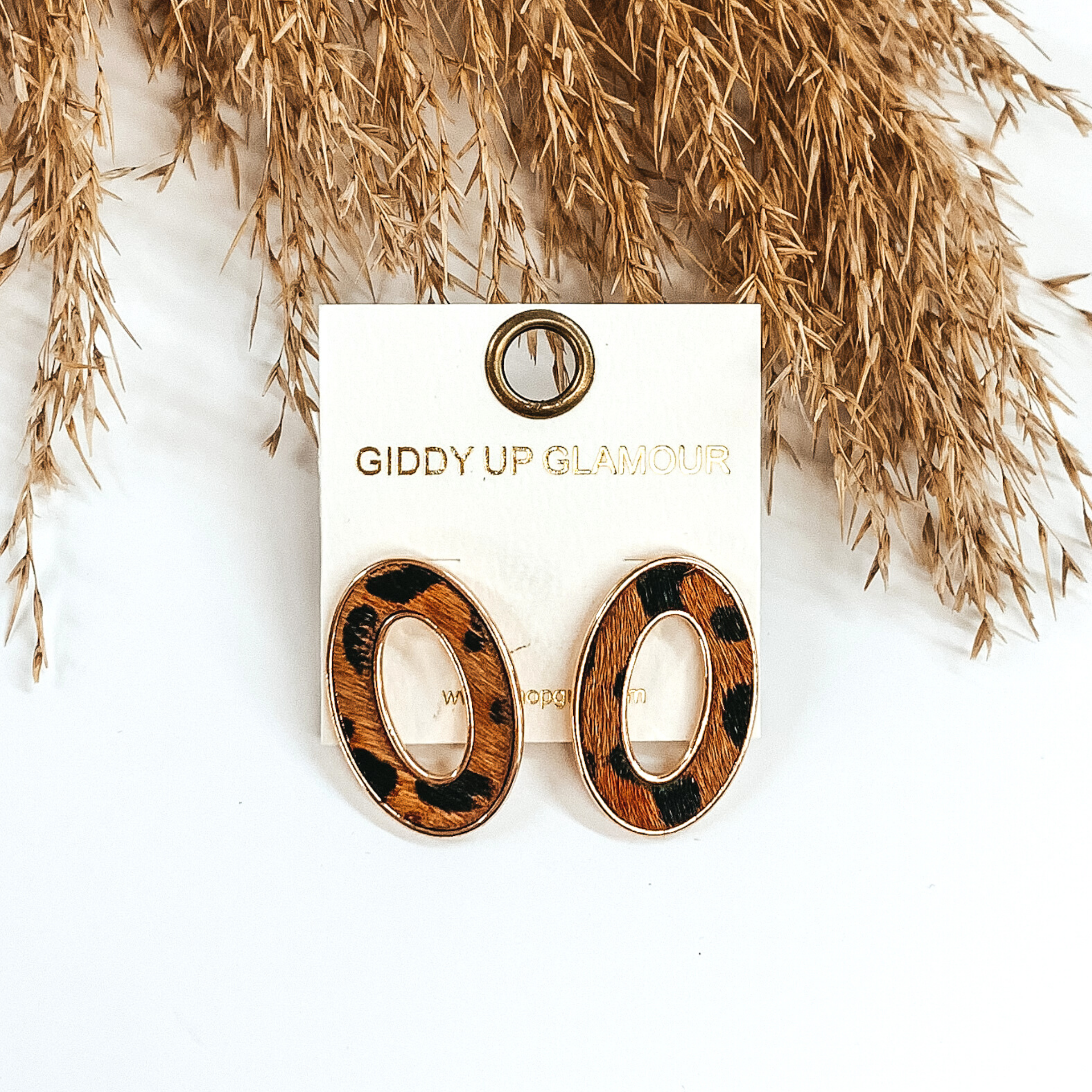 Gold, open, oval stud earrings with a brown hide inlay that has a black animal print. These earrings are pictured laying on top of tan floral on a white background. 