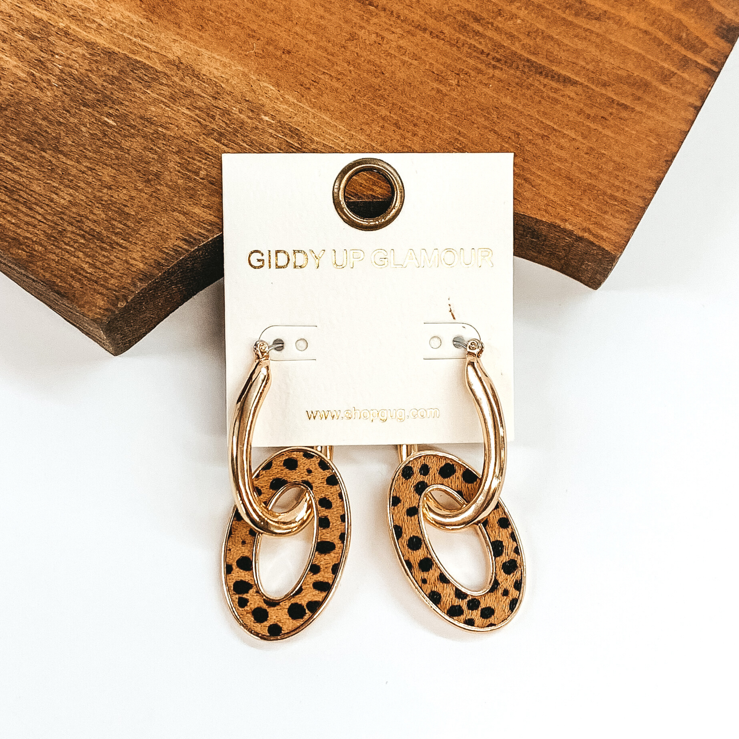 Gold oval hoop earrings with another oval link hanging. The hanging oval has a brown hide inlay that has a black dotted print. These earrings are pictured laying against some brown wood on a white background.