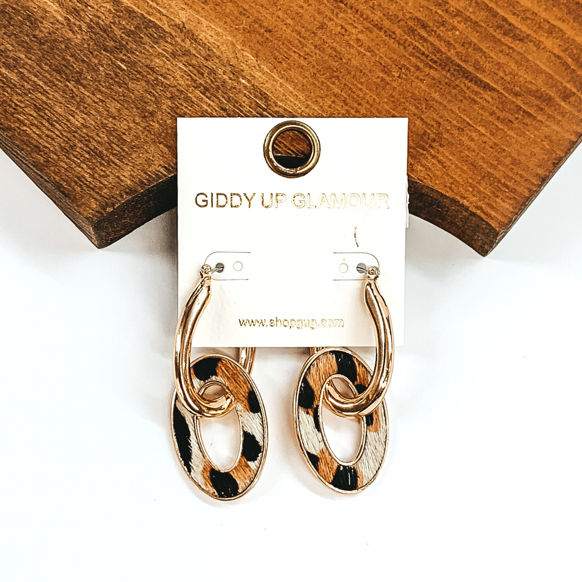 Gold oval hoop earrings with another oval link hanging. The hanging oval has a white hide inlay that has a black and brown animal print. These earrings are pictured laying against some brown wood on a white background.  