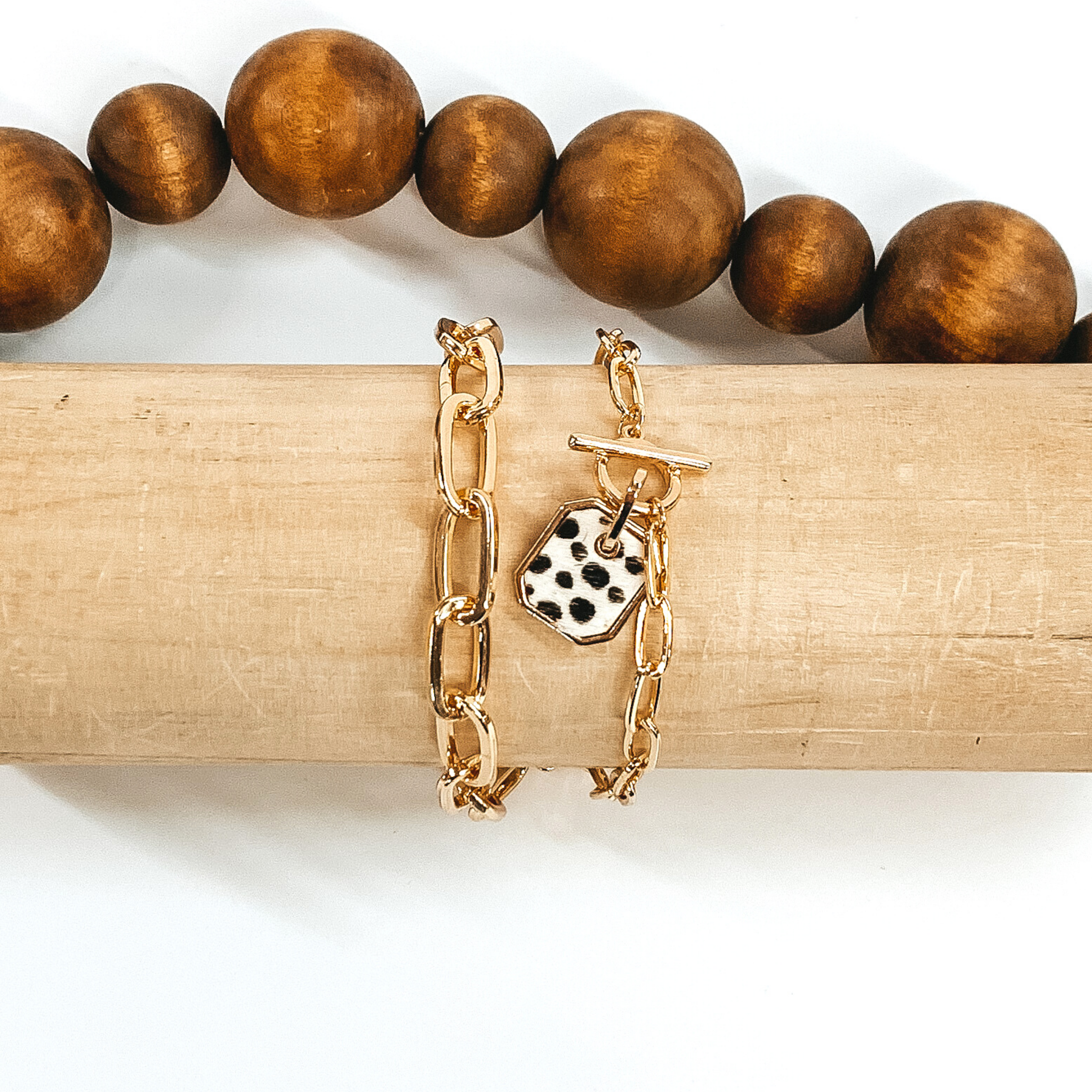 Gold two chained bracelet with an octagon pendant with a white hide inlay with a black dotted print. this bracelet is pictured on a light tan bracelet holder on a white background with brown beads in the background. 