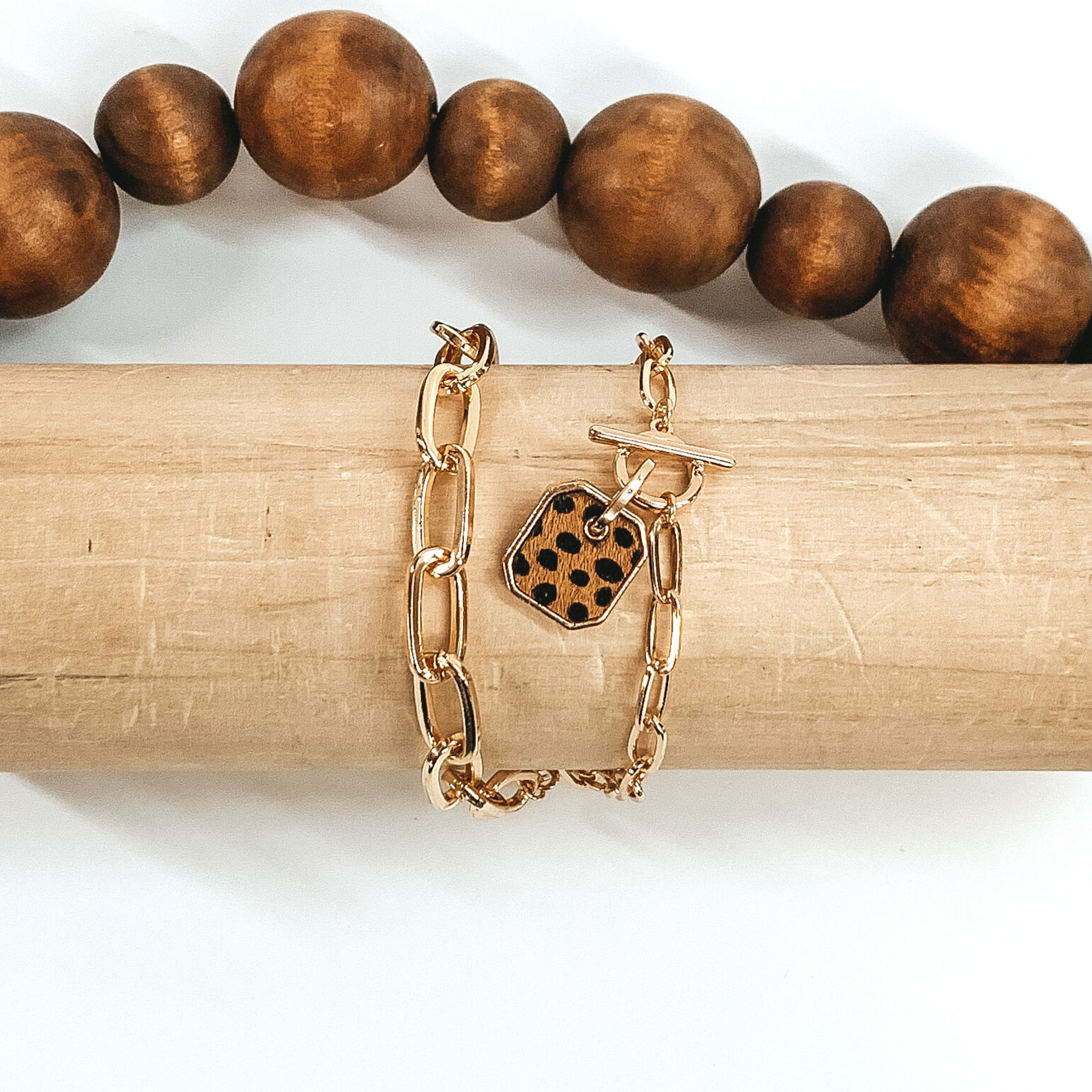 Gold two chained bracelet with an octagon pendant with a brown hide inlay with a black dotted print. this bracelet is pictured on a light tan bracelet holder on a white background with brown beads in the background. 