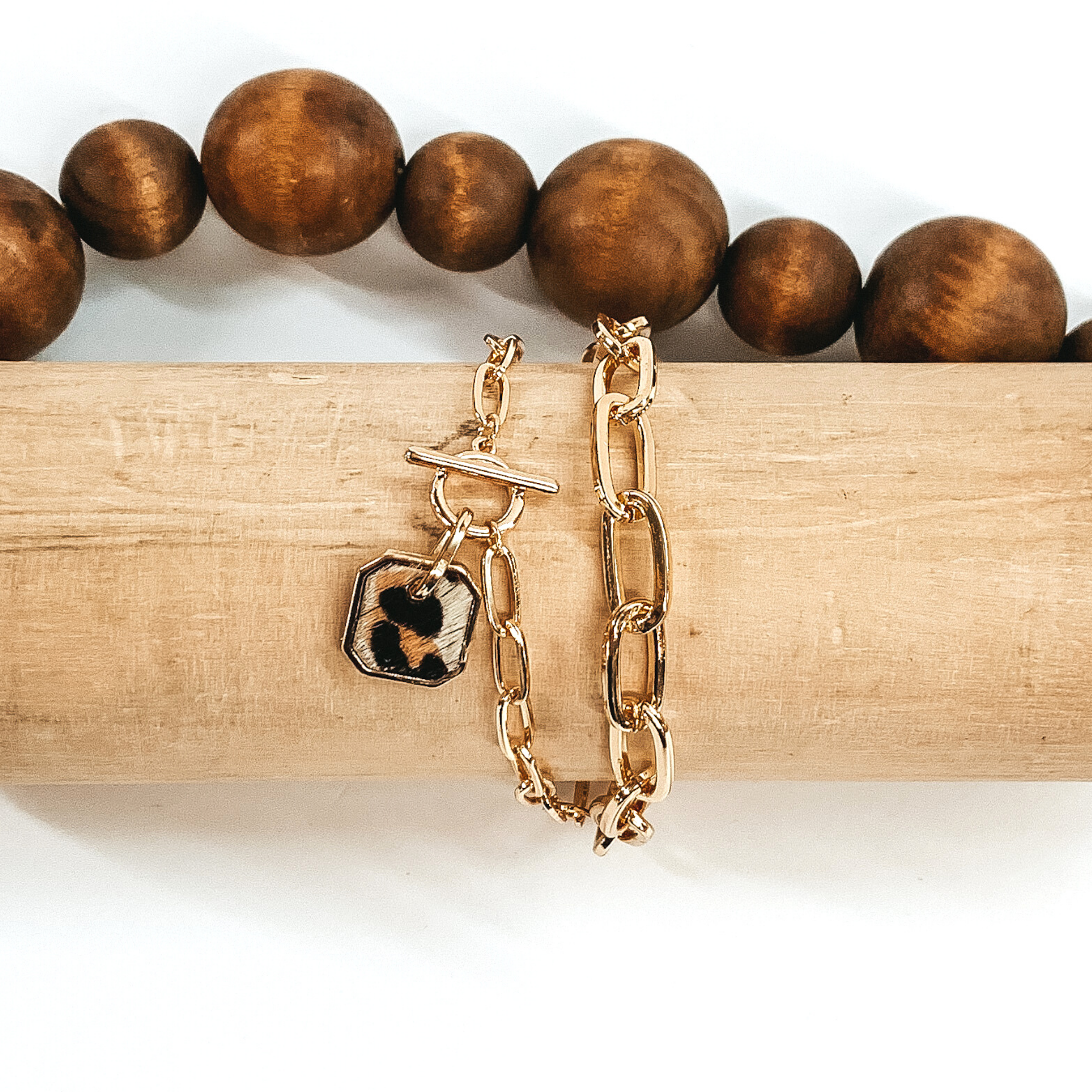 Gold two chained bracelet with an octagon pendant with a white hide inlay with a black and brown animal print. This bracelet is pictured on a light tan bracelet holder on a white background with brown beads in the background. 