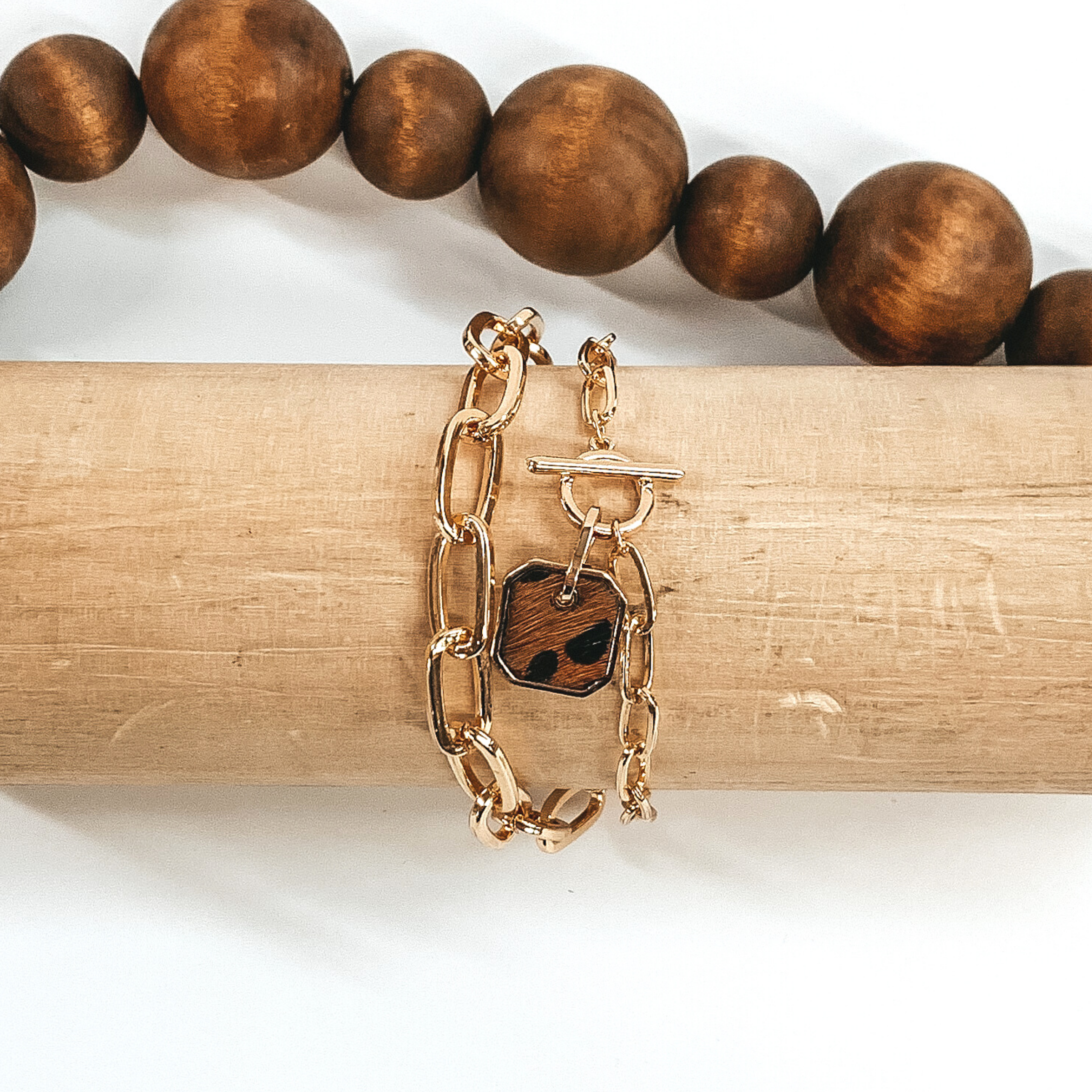 Gold two chained bracelet with an octagon pendant with a brown hide inlay with a black animal print. This bracelet is pictured on a light tan bracelet holder on a white background with brown beads in the background.