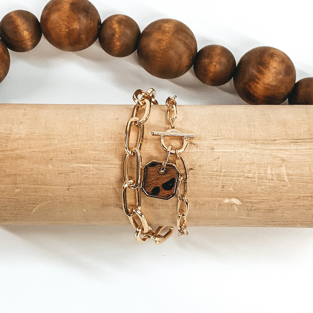 Gold two chained bracelet with an octagon pendant with a brown hide inlay with a black animal print. This bracelet is pictured on a light tan bracelet holder on a white background with brown beads in the background.
