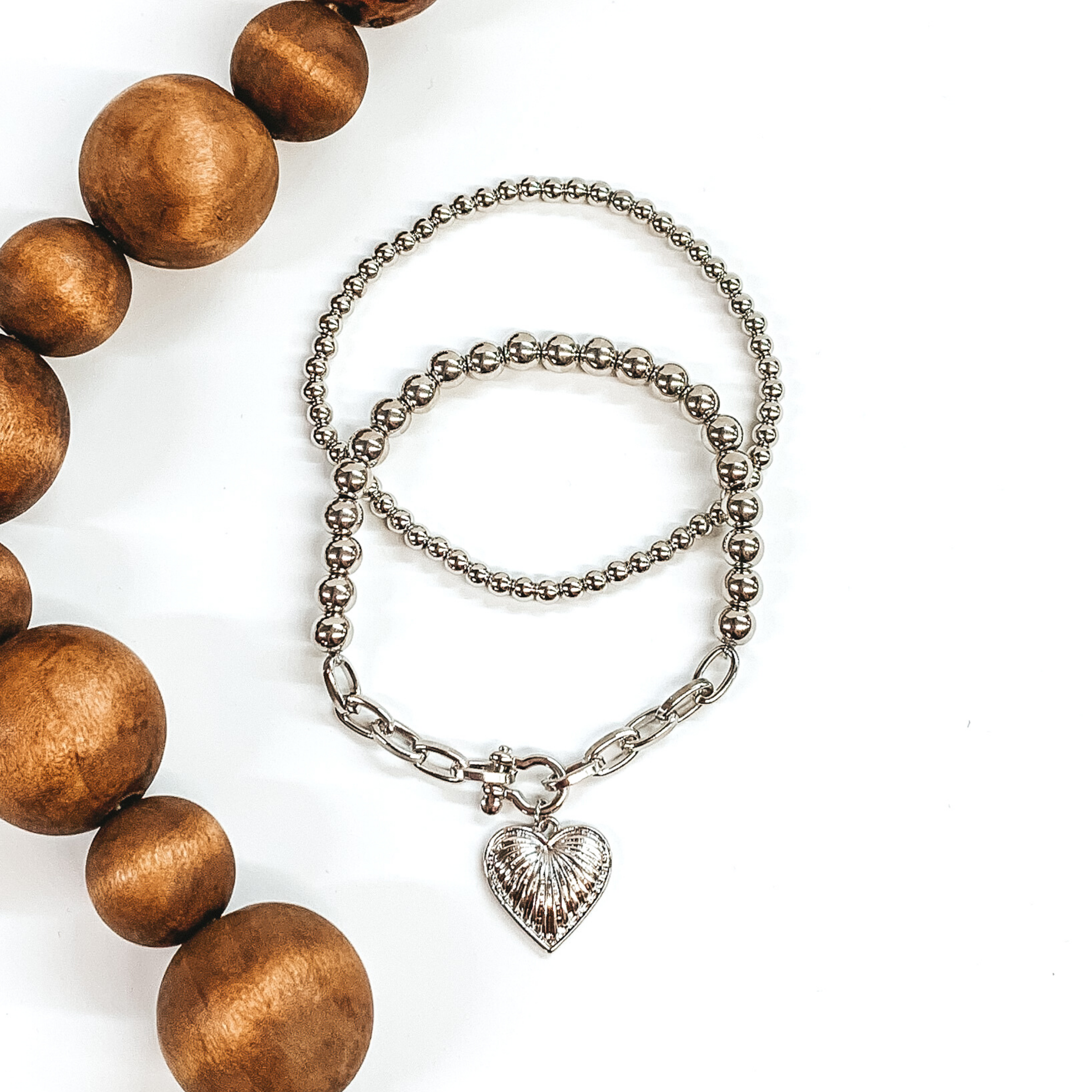  This silver bracelet set includes two beaded bracelets. One is a plain beaded bracelet and the other one has a chained segment with heart charm. This bracelet set is pictured on a white background with brown beads on the side of the picture.