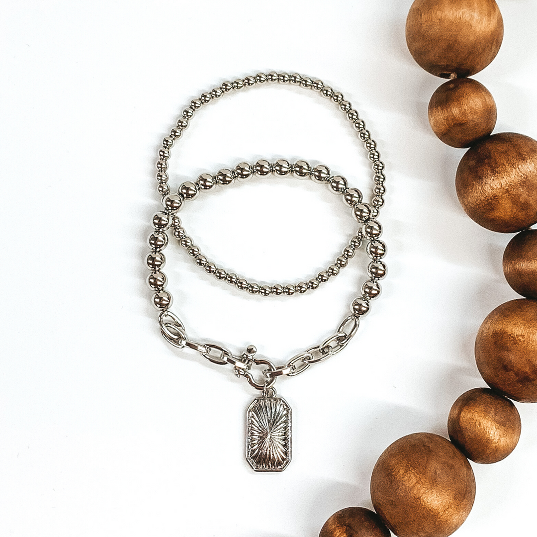 This silver bracelet set includes two beaded bracelets. One is a plain beaded bracelet and the other one has a chained segment with rectangle charm. This bracelet set is pictured on a white background with brown beads on the side of the picture.