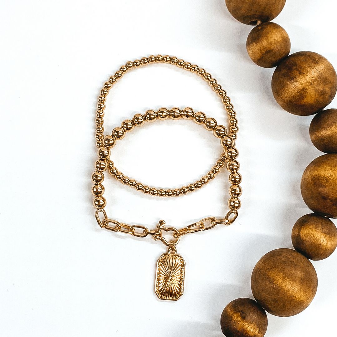 This gold bracelet set includes two beaded bracelets. One is a plain beaded bracelet and the other one has a chained segment with rectangle charm. This bracelet set is pictured on a white background with brown beads on the side of the picture.