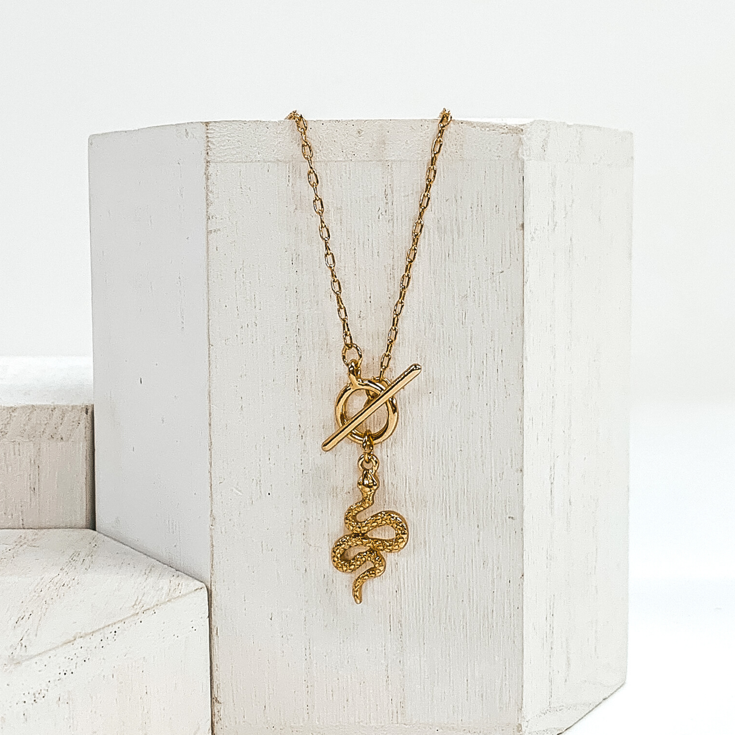 This is a gold necklace that has a front toggle clasp and a gold snake charm. This necklace is pictured laying on a white block and on a white background with other white blocks around it.