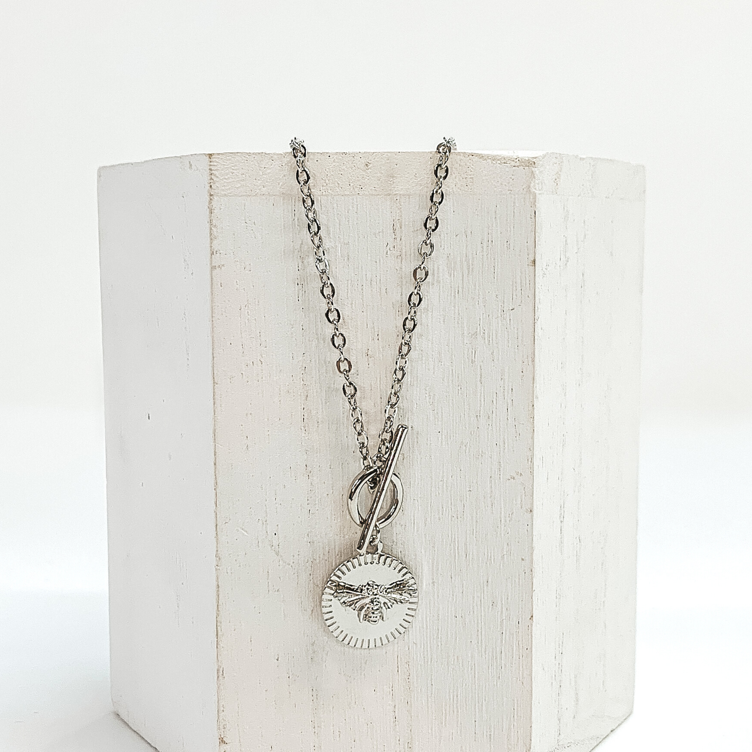 This is a silver necklace that has a front toggle clasp and a circle pendant with a bumble bee charm. This necklace is pictured laying on a white block and on a white background. 