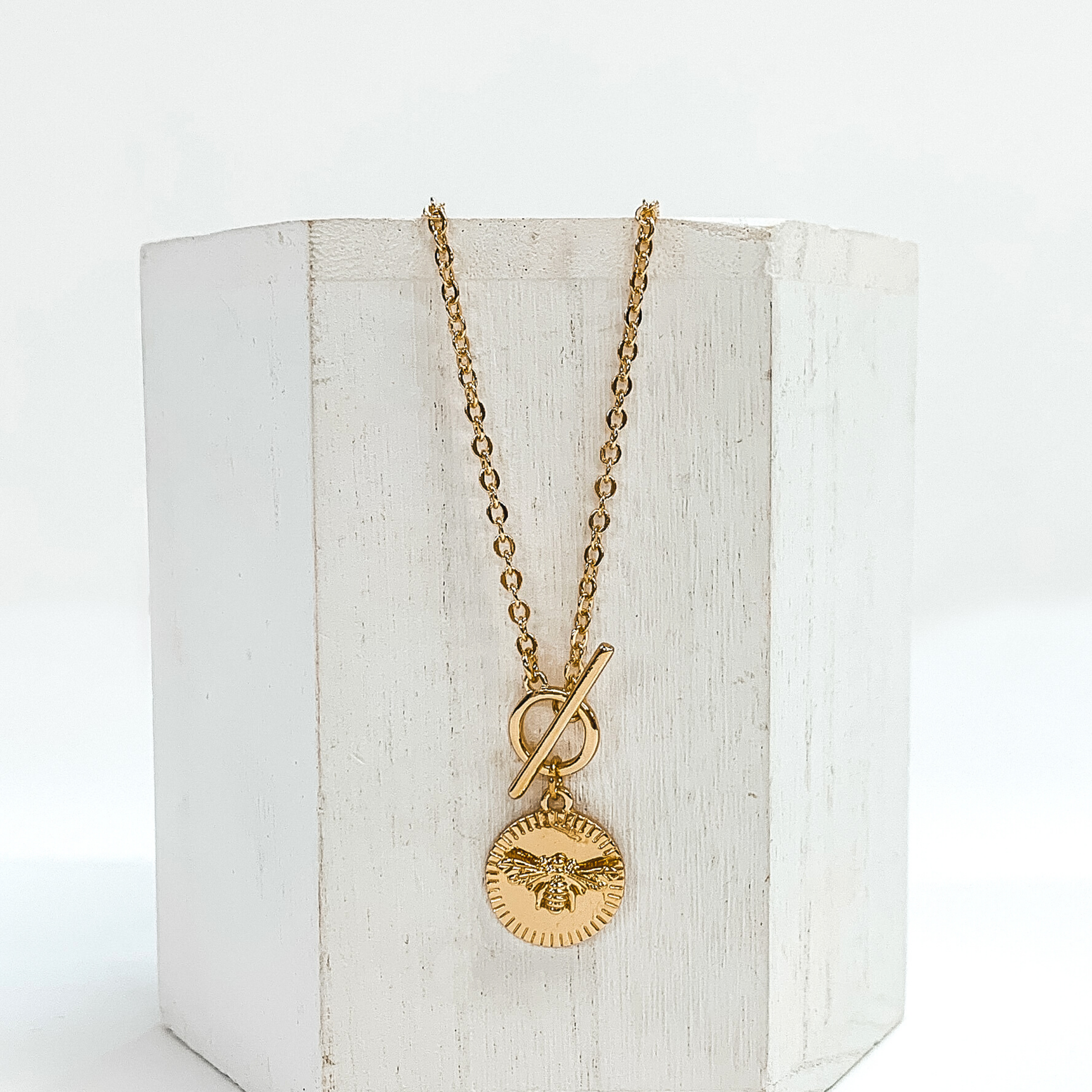 This is a gold necklace that has a front toggle clasp and a circle pendant with a bumble bee charm. This necklace is pictured laying on a white block and on a white background.
