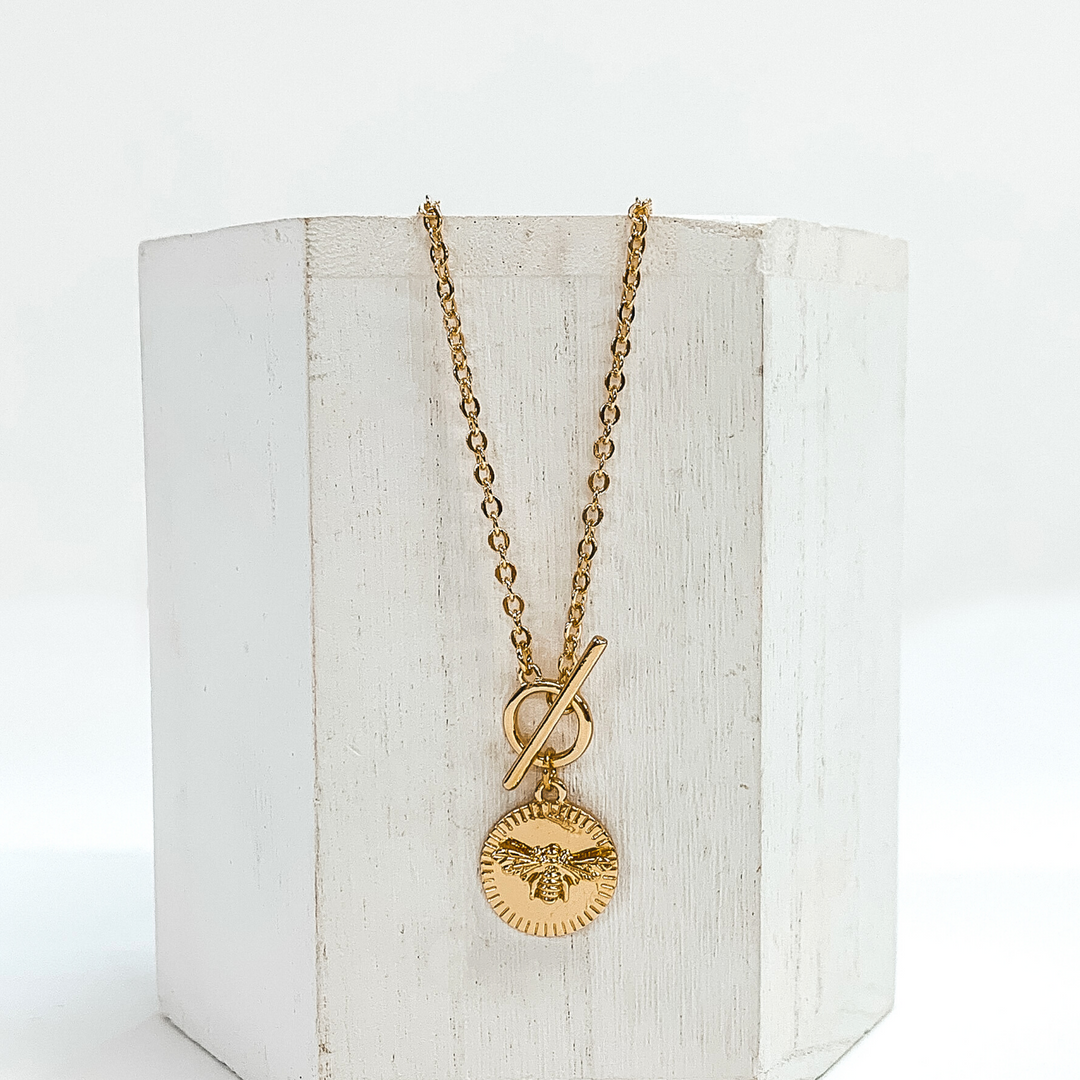 This is a gold necklace that has a front toggle clasp and a circle pendant with a bumble bee charm. This necklace is pictured laying on a white block and on a white background.
