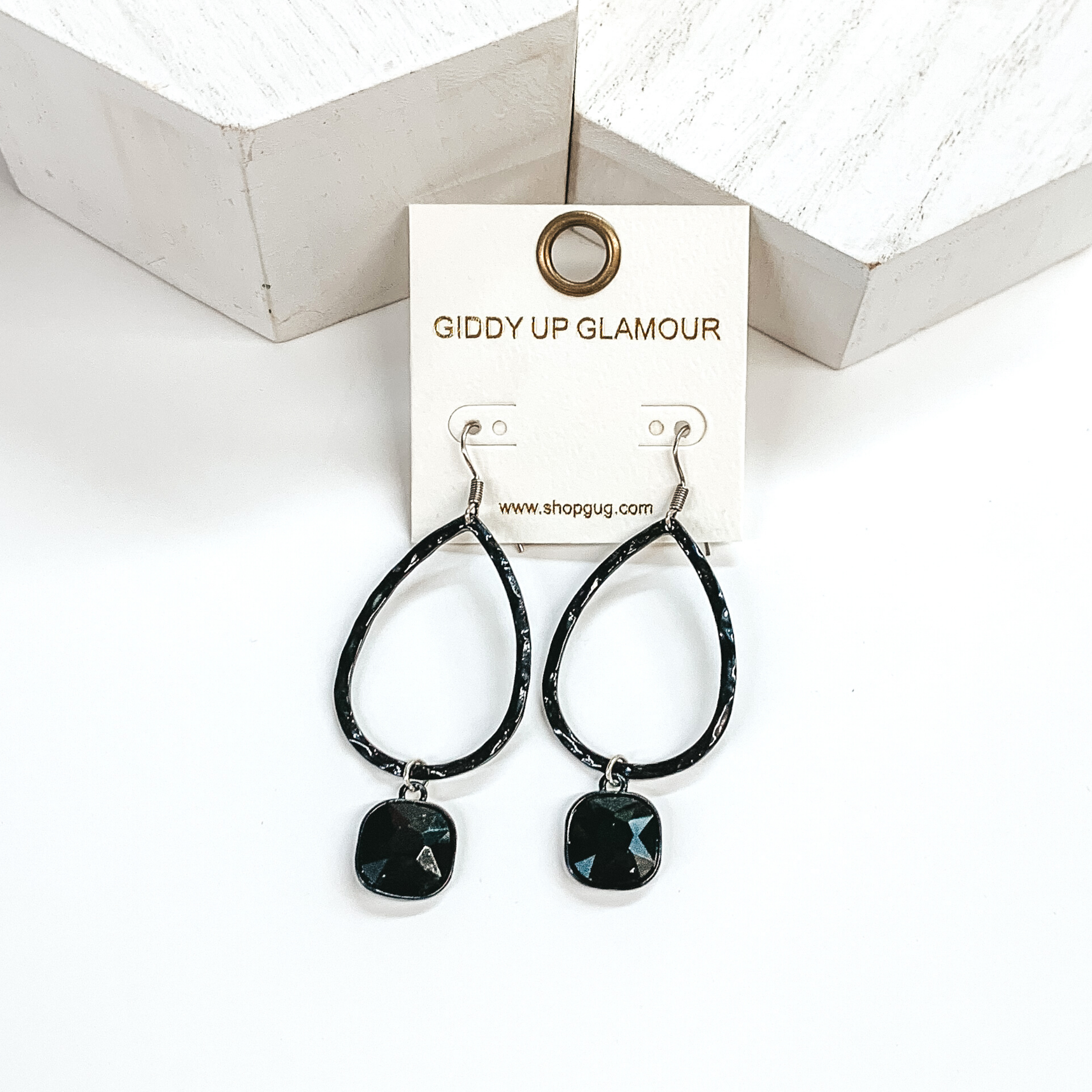 Black teardrop hammered earrings with a hanging cushion cut black crystal. These earrings are pictured on a white background with white blocks at the top of the picture. 