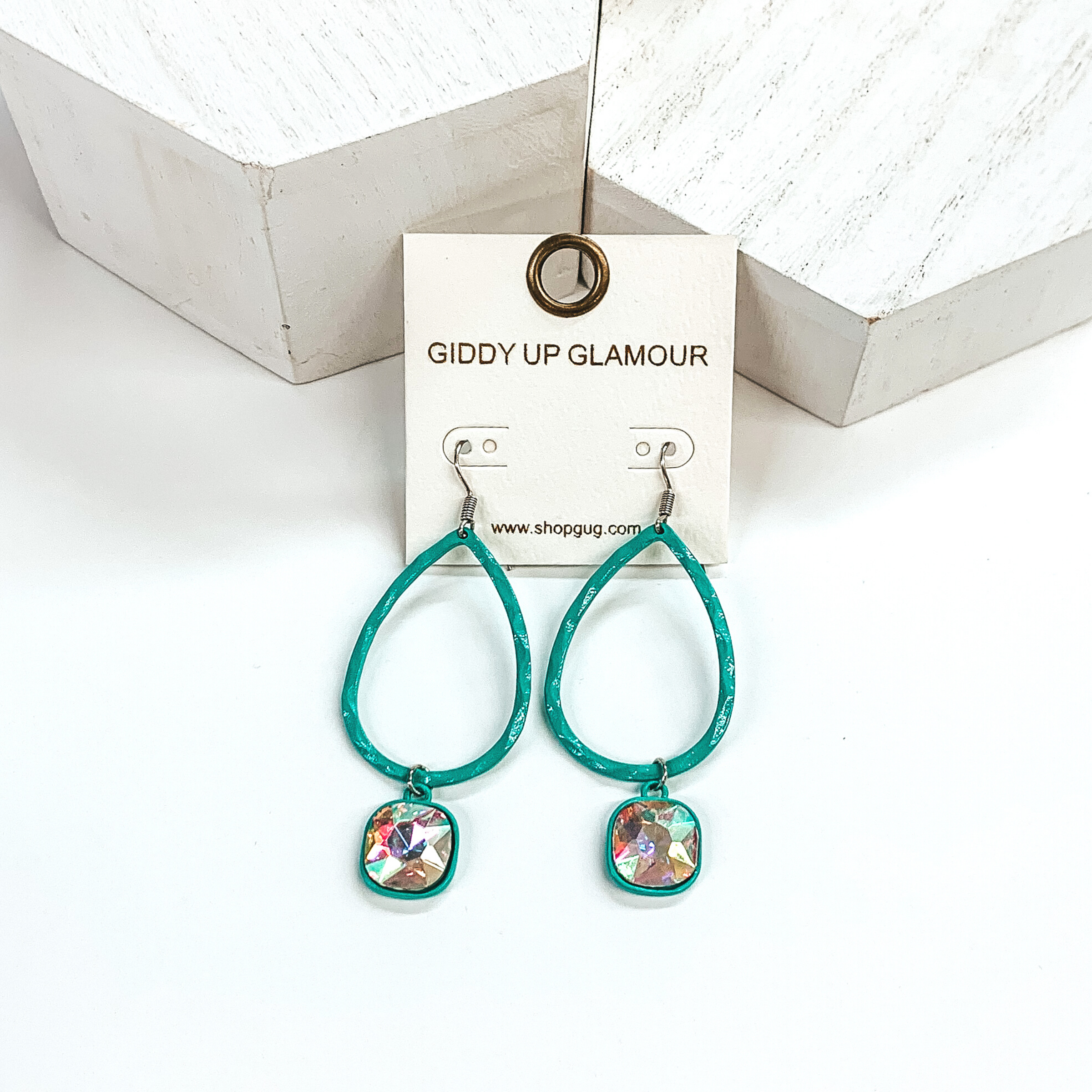 Turquoise teardrop hammered earrings with a hanging cushion cut ab crystal. These earrings are pictured on a white background with white blocks at the top of the picture. 