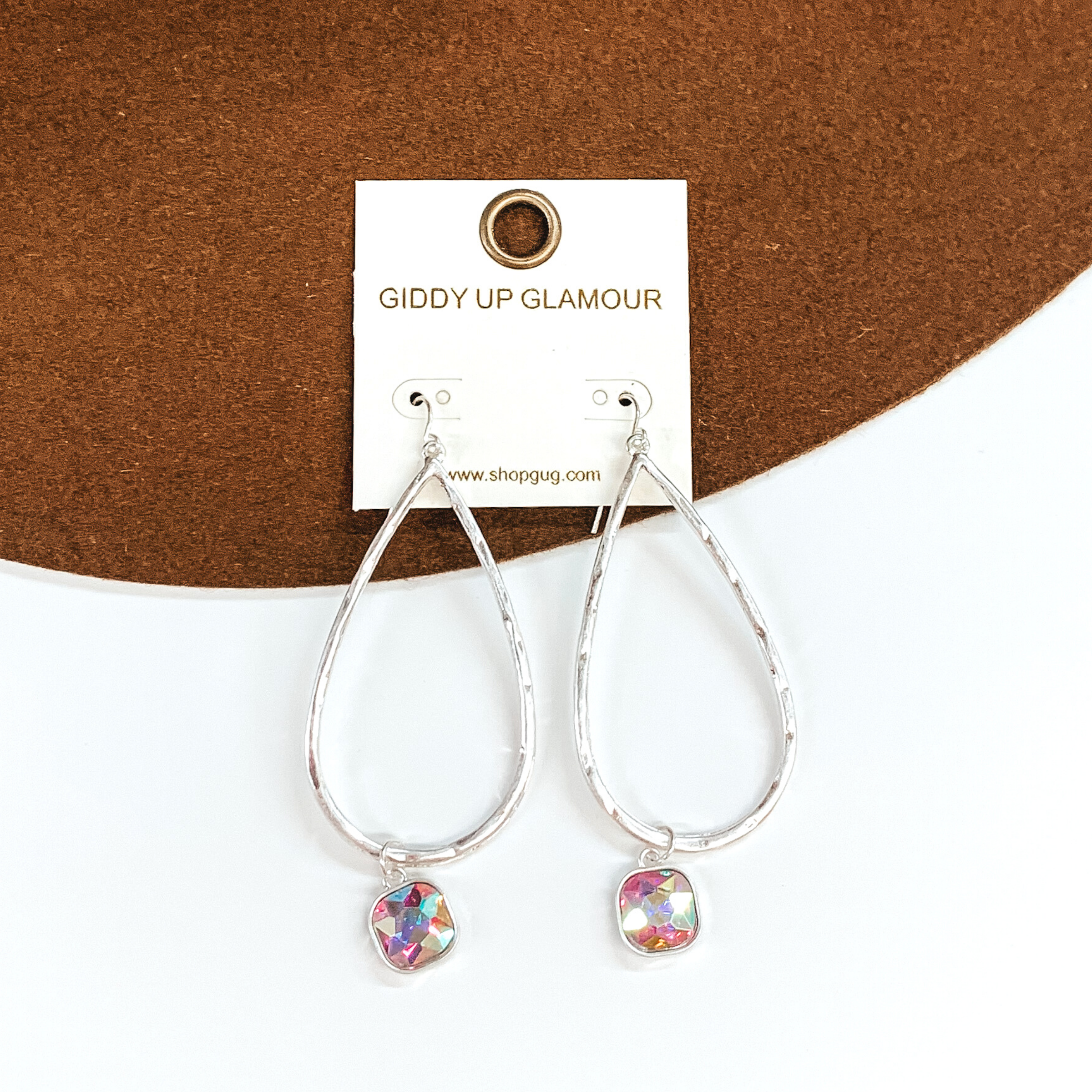 Silver teardrop hammered earrings with a hanging cushion cut ab crystal. These earrings are pictured on a white and brown background. 
